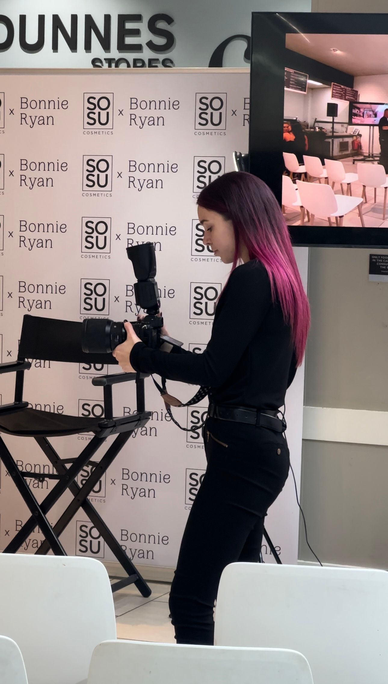 Woman with pink hair holding a camera at a Bonnie Ryan SOSU Cosmetics branded event backdrop.