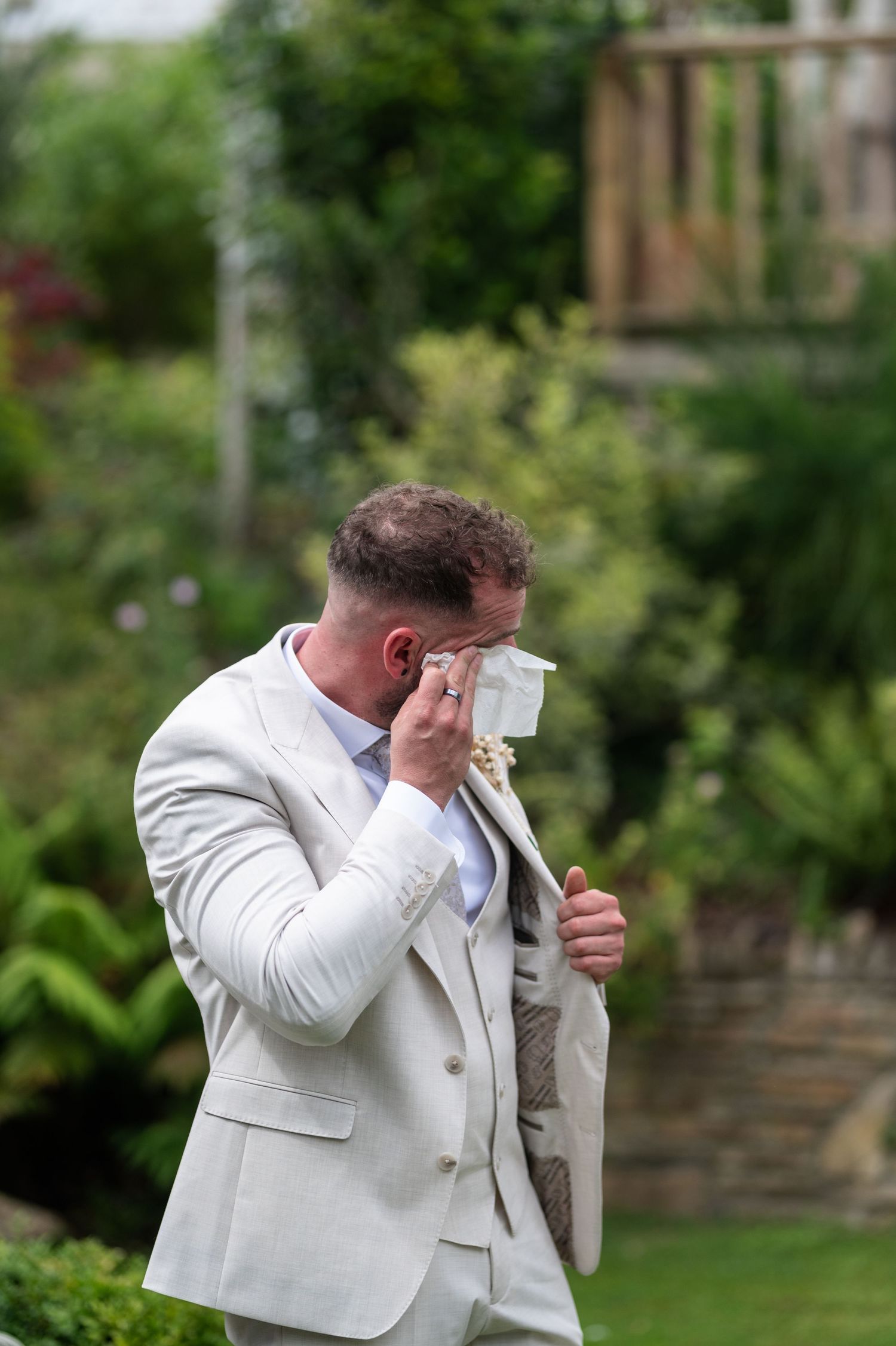 Emotional groom in cream suit wiping tears during outdoor garden wedding ceremony.