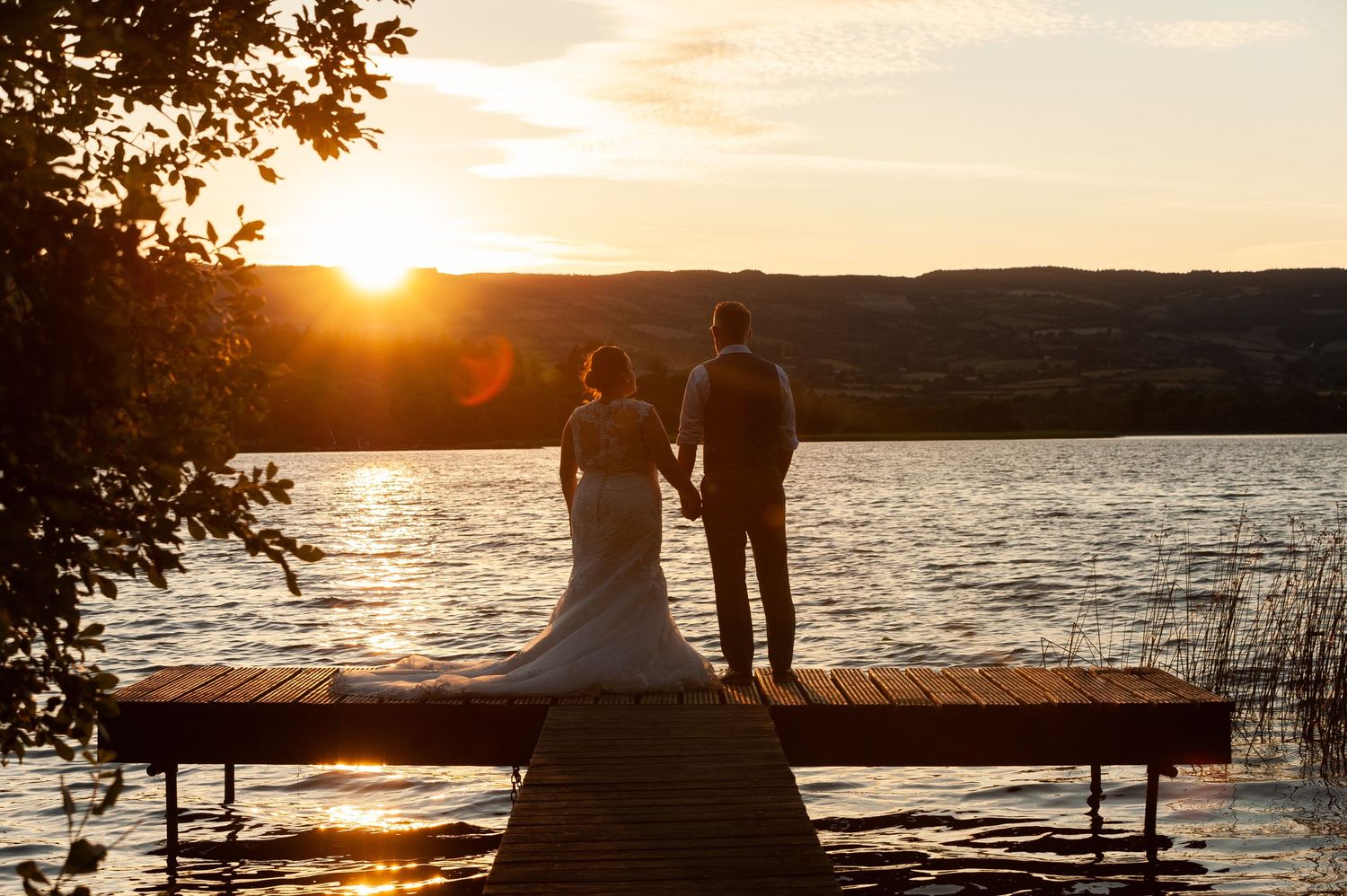 Bride and groom holding hands on a wooden dock at sunset overlooking a serene lake with golden reflections.