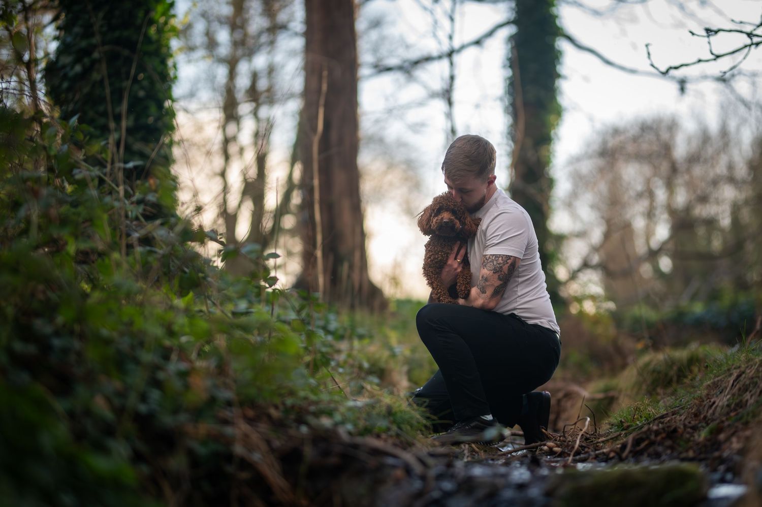 Man crouching in woodland setting, tenderly embracing a small brown dog among green foliage and tall trees at dusk.