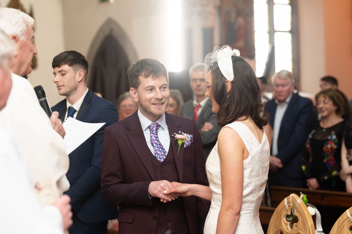 Groom in burgundy suit smiling at bride during wedding ceremony in a church.