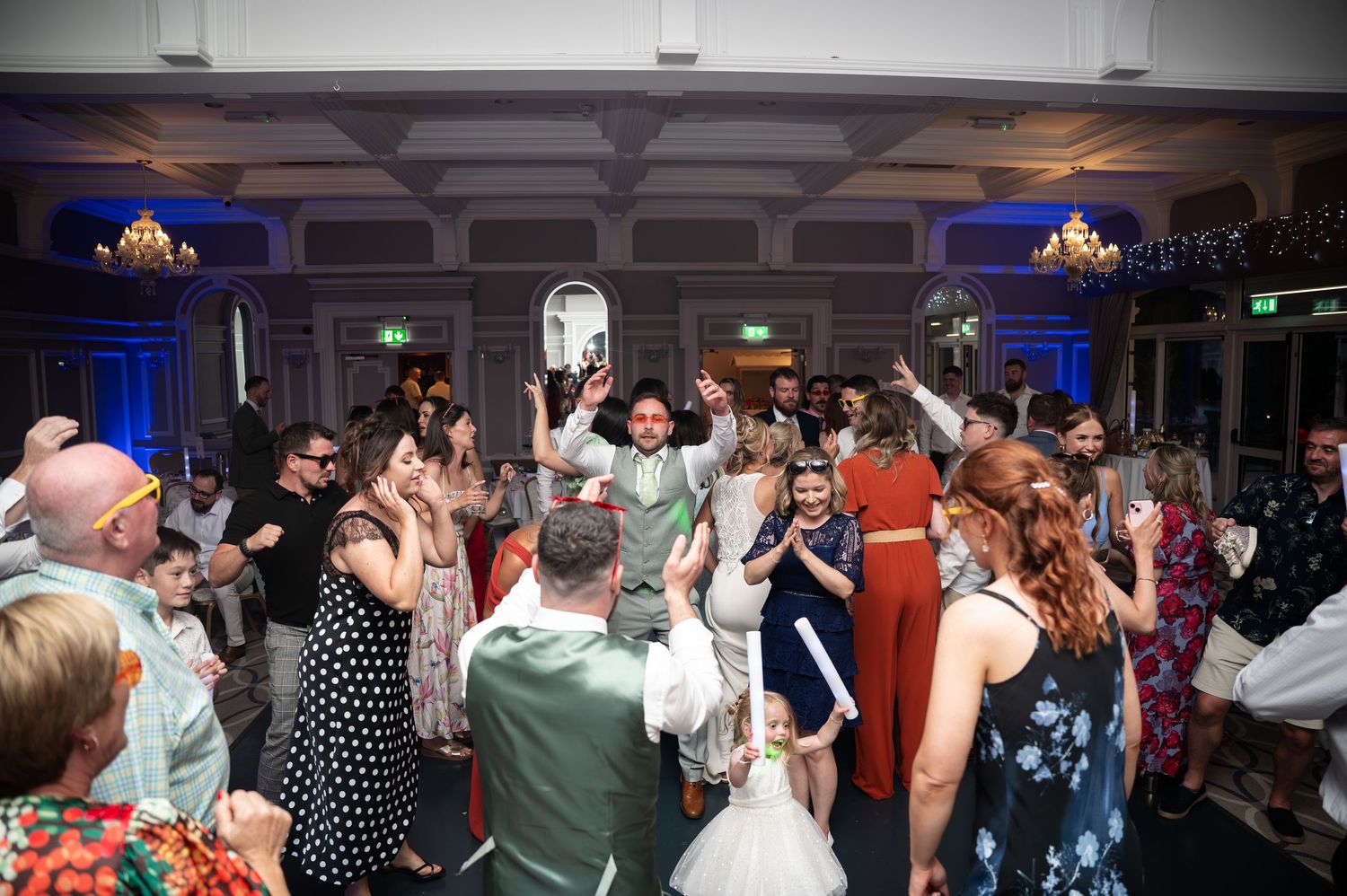 Lively wedding reception dance floor packed with celebrating guests under blue ballroom lighting.