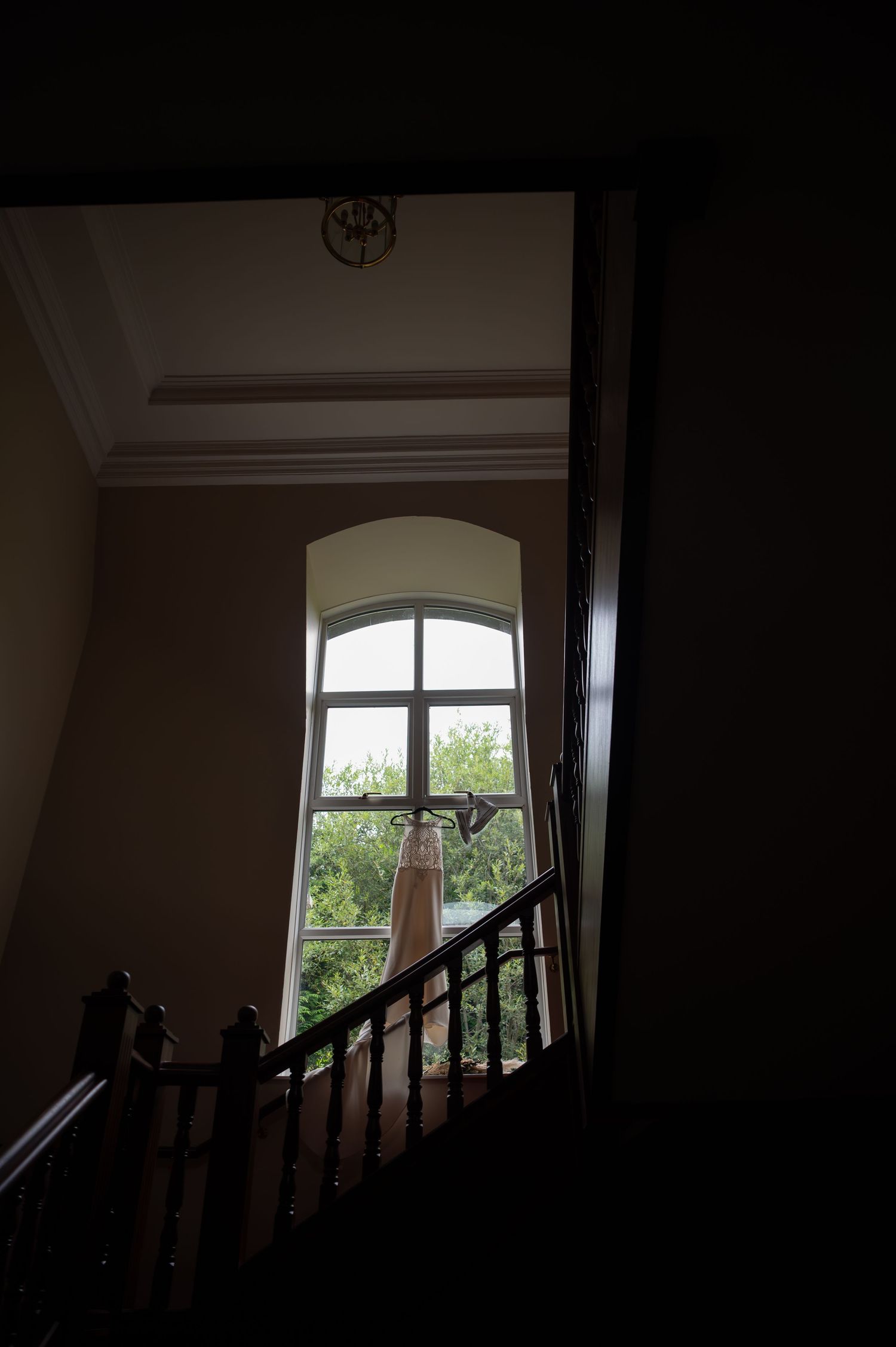A wedding dress hangs near an arched window on a dark wooden staircase in an elegant venue.