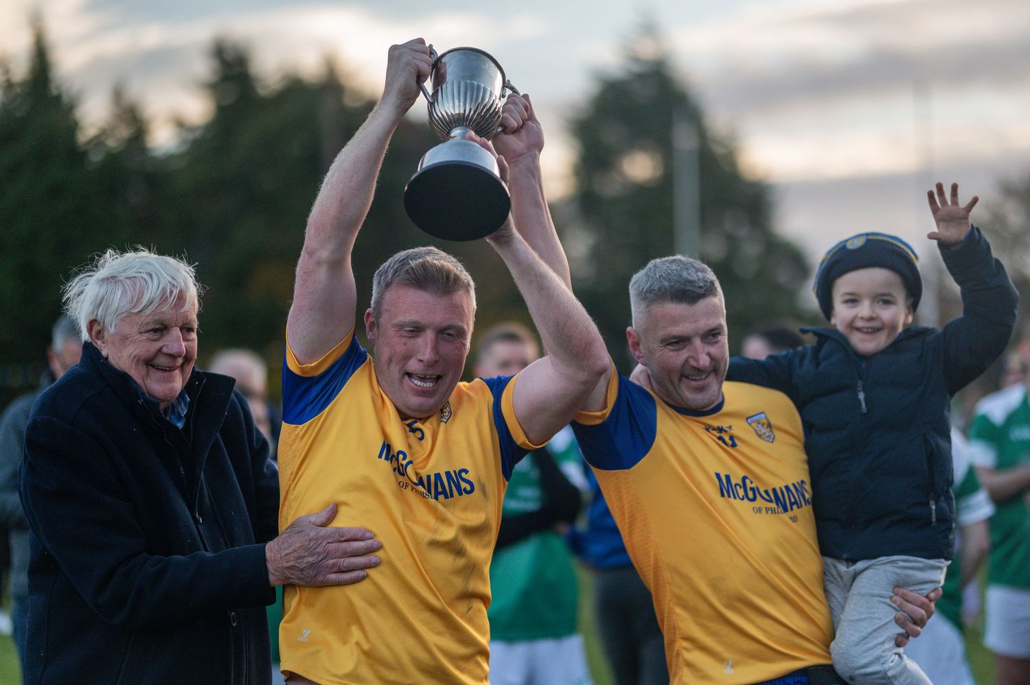 Two men in yellow and blue jerseys celebrate a trophy win with an elderly woman and a child at a sports event.
