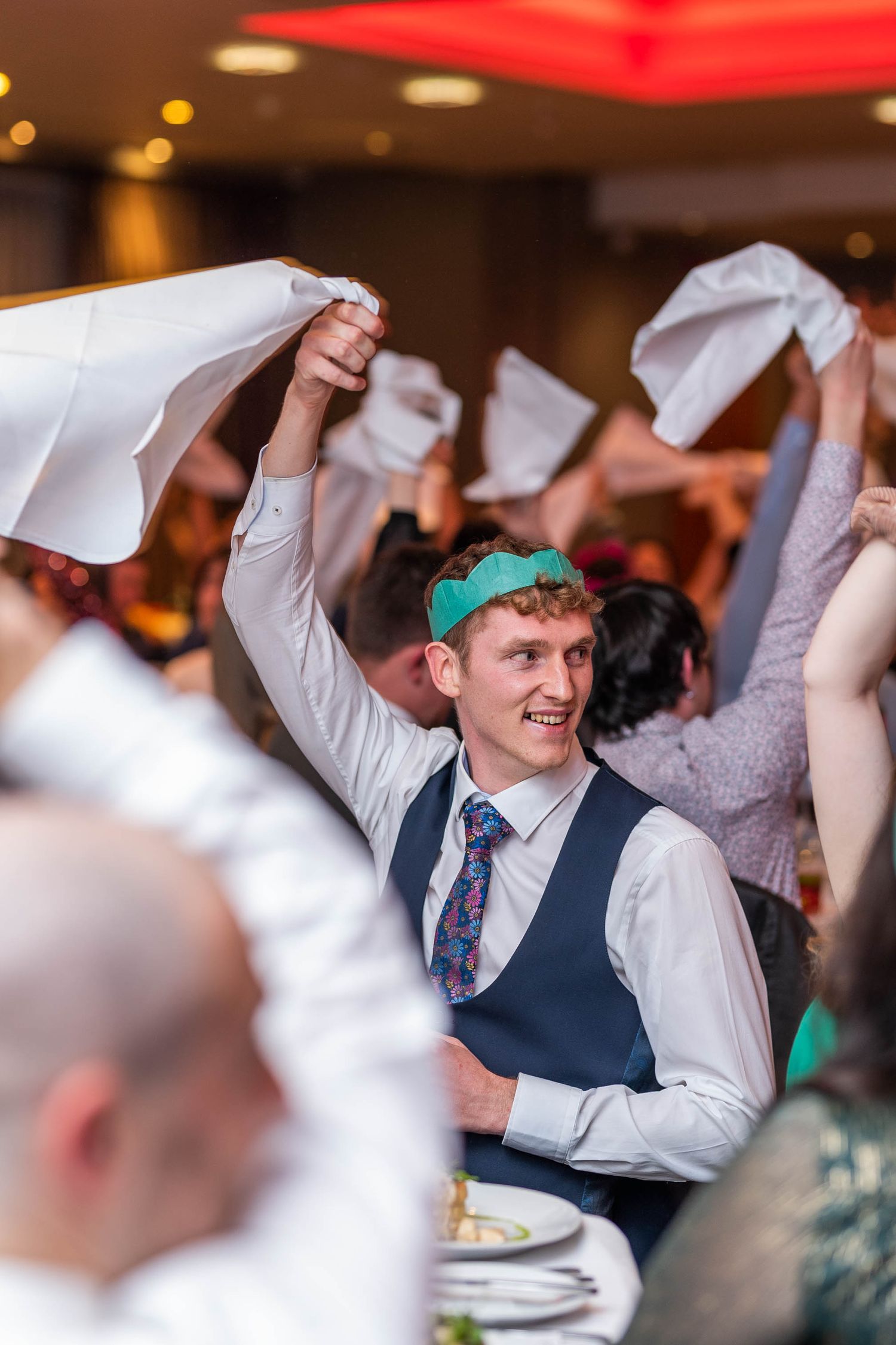 Man in waistcoat waving napkin at a lively formal dinner party celebration.