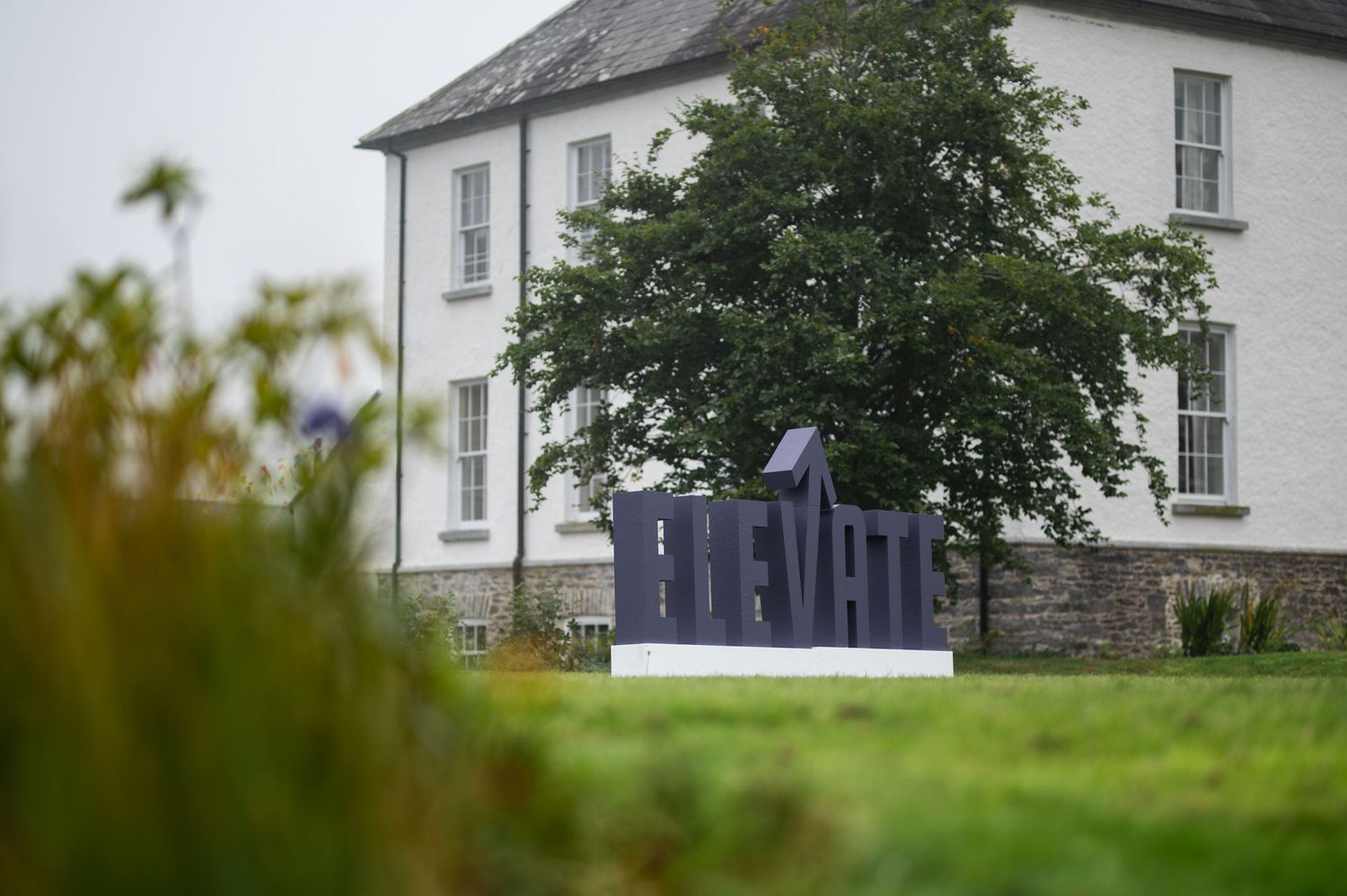 Sculpture with bold letters on green lawn before a white Georgian building and tree in overcast Ireland.