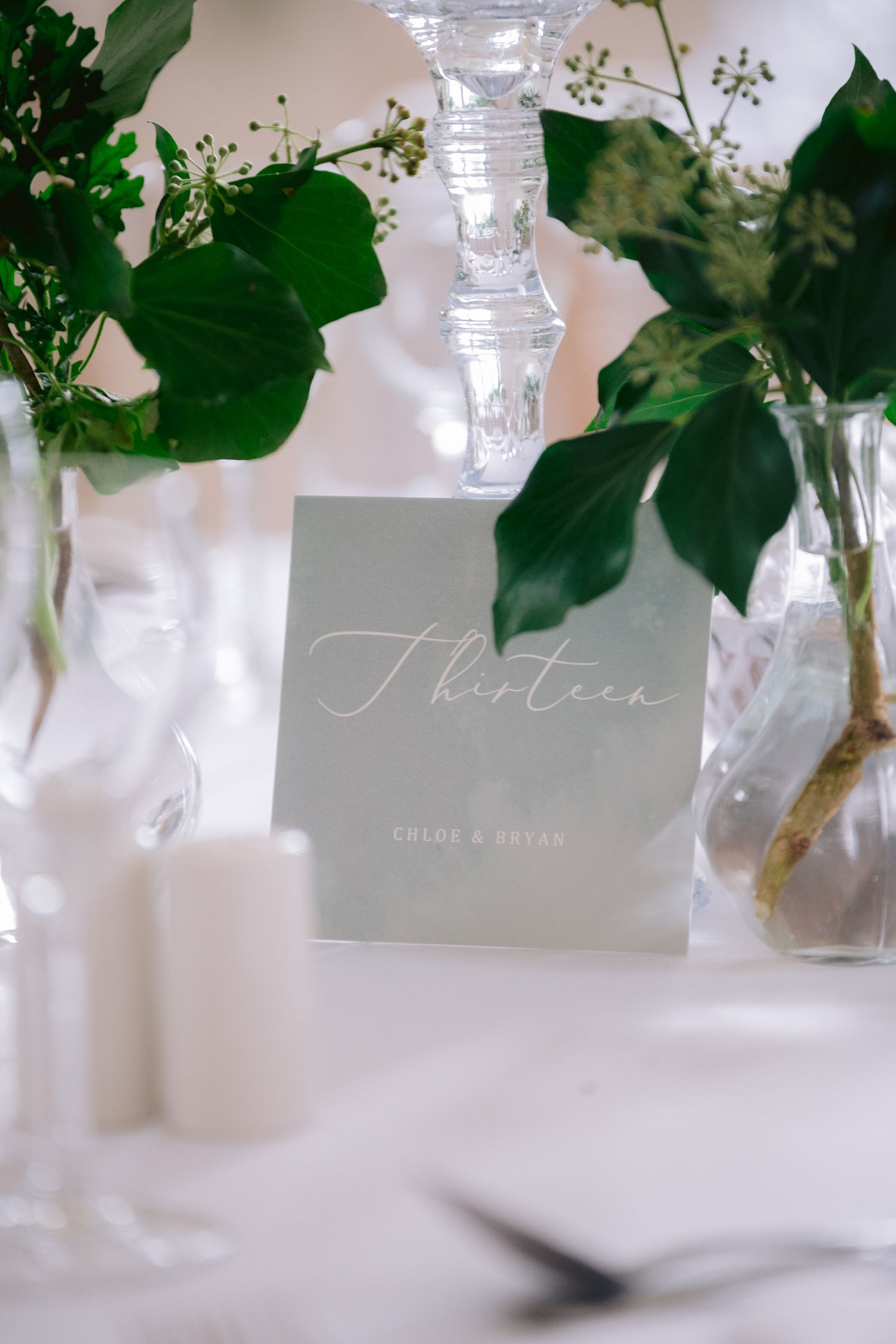 Elegant wedding table number thirteen card with sage green design, surrounded by lush greenery and white candles.