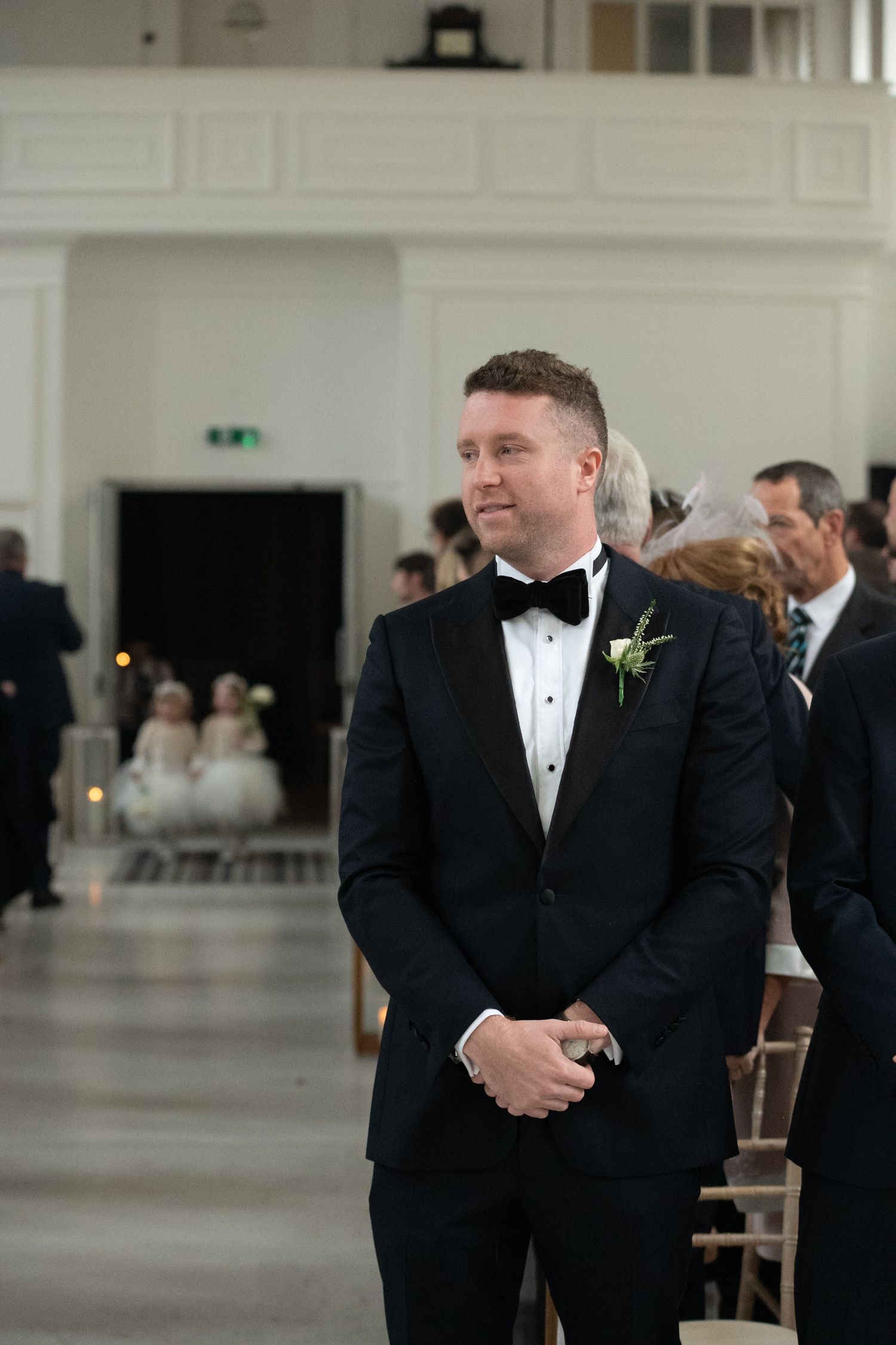 Groom in black tuxedo with bow tie and boutonniere stands at altar awaiting bride during wedding ceremony.