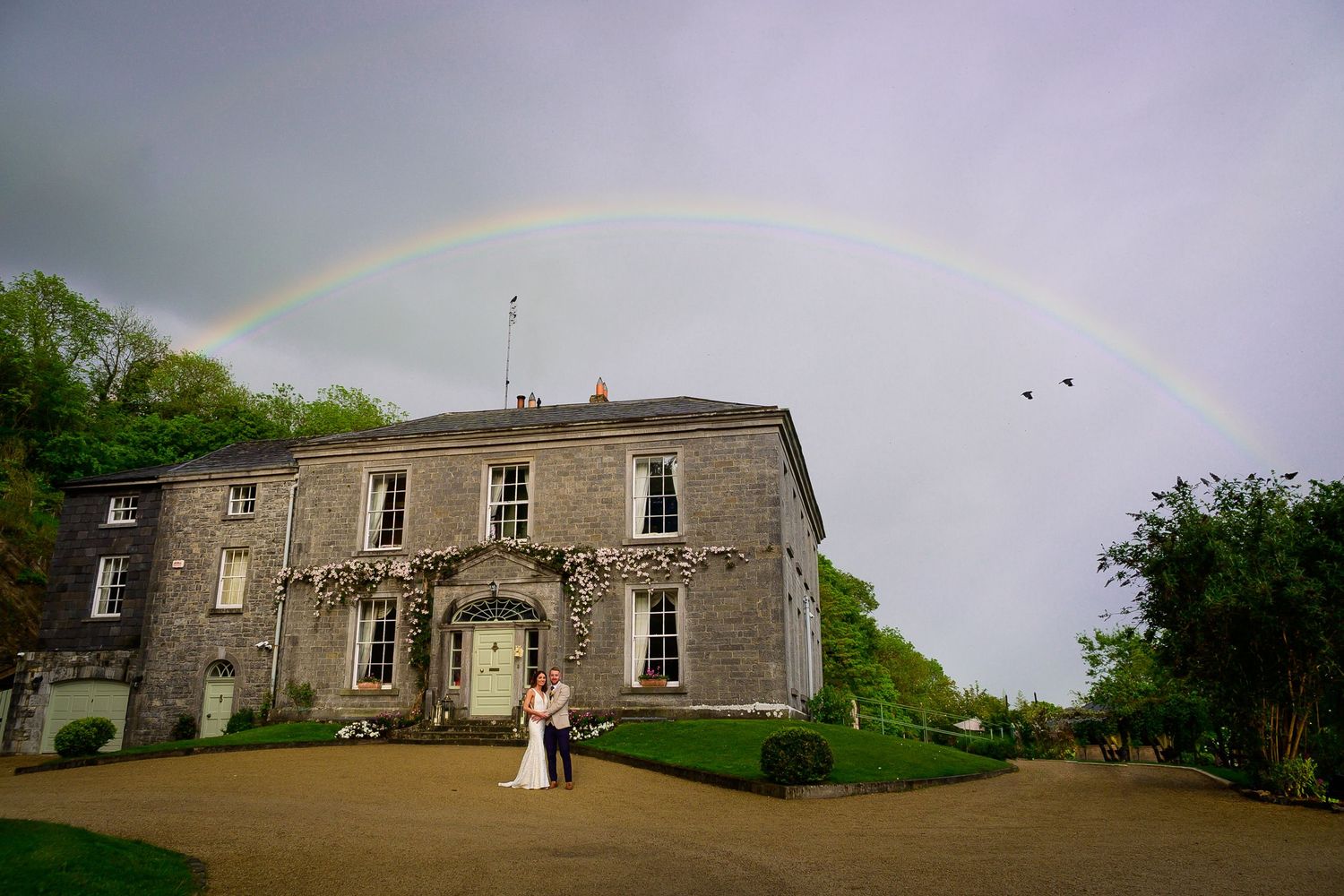 Wedding couple stands before a grand stone manor house under a stunning full rainbow in Ireland.