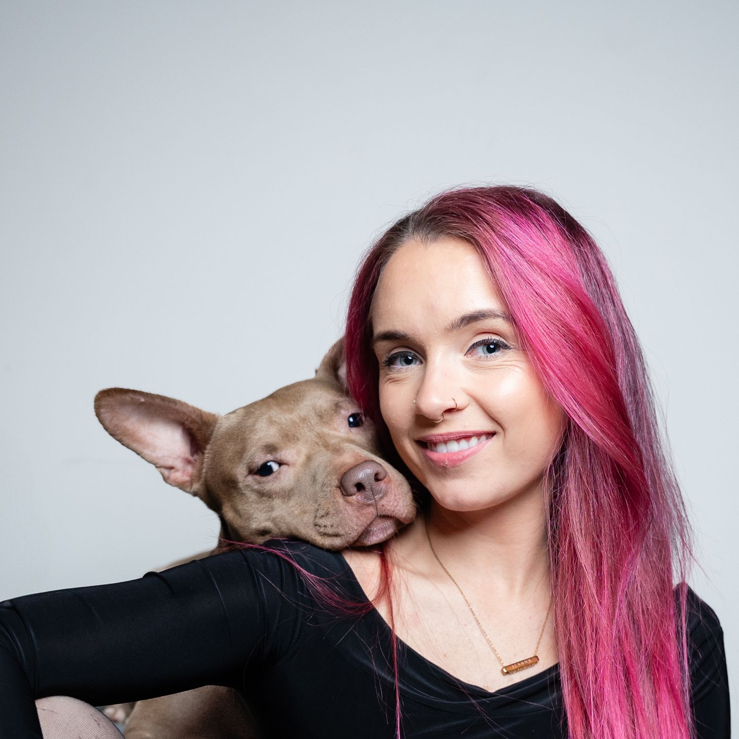 Smiling young woman with pink hair holding a brown pit bull puppy against a gray background.