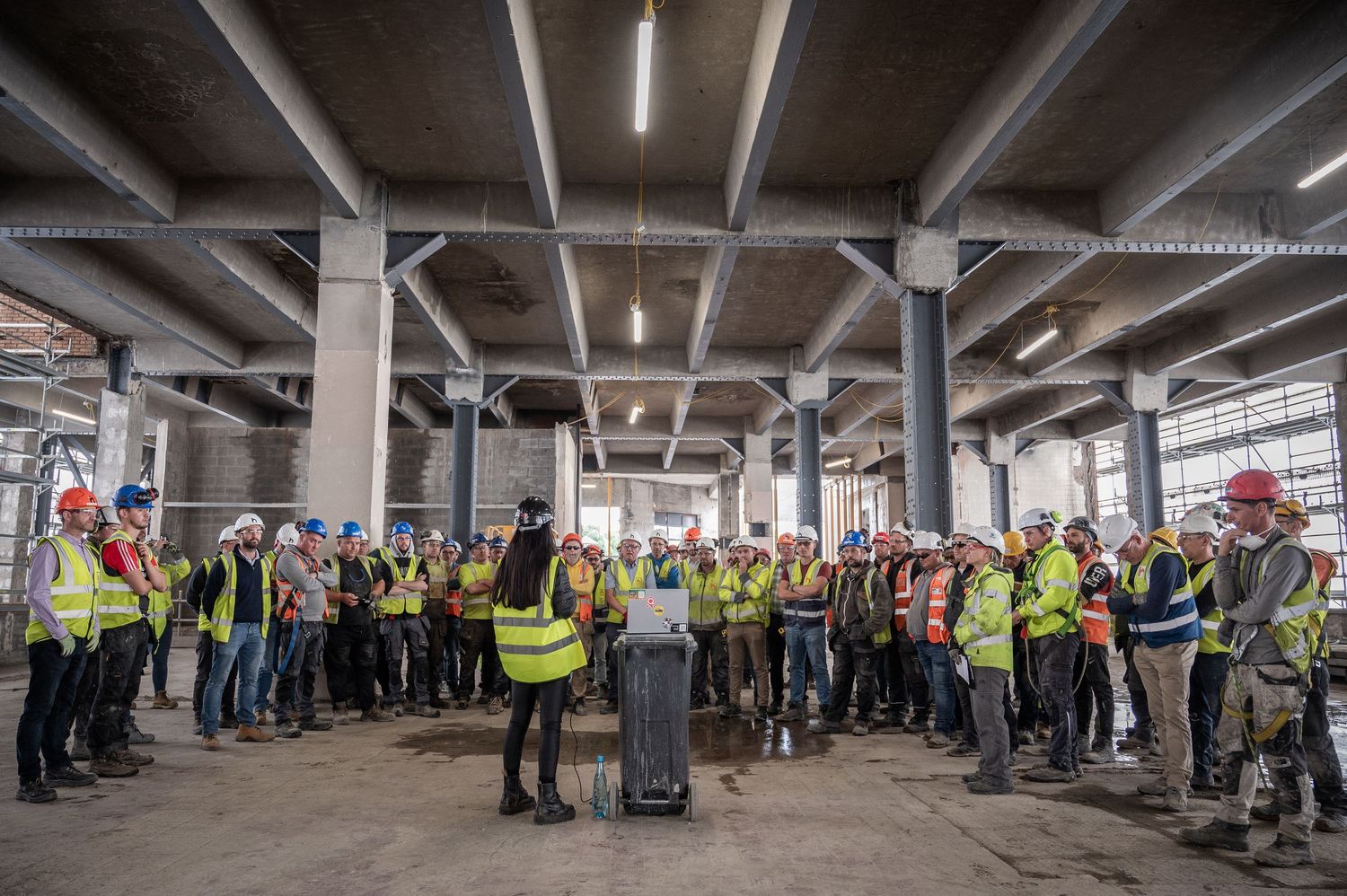 Large group of workers in hi-vis vests attending a safety briefing inside a construction site.