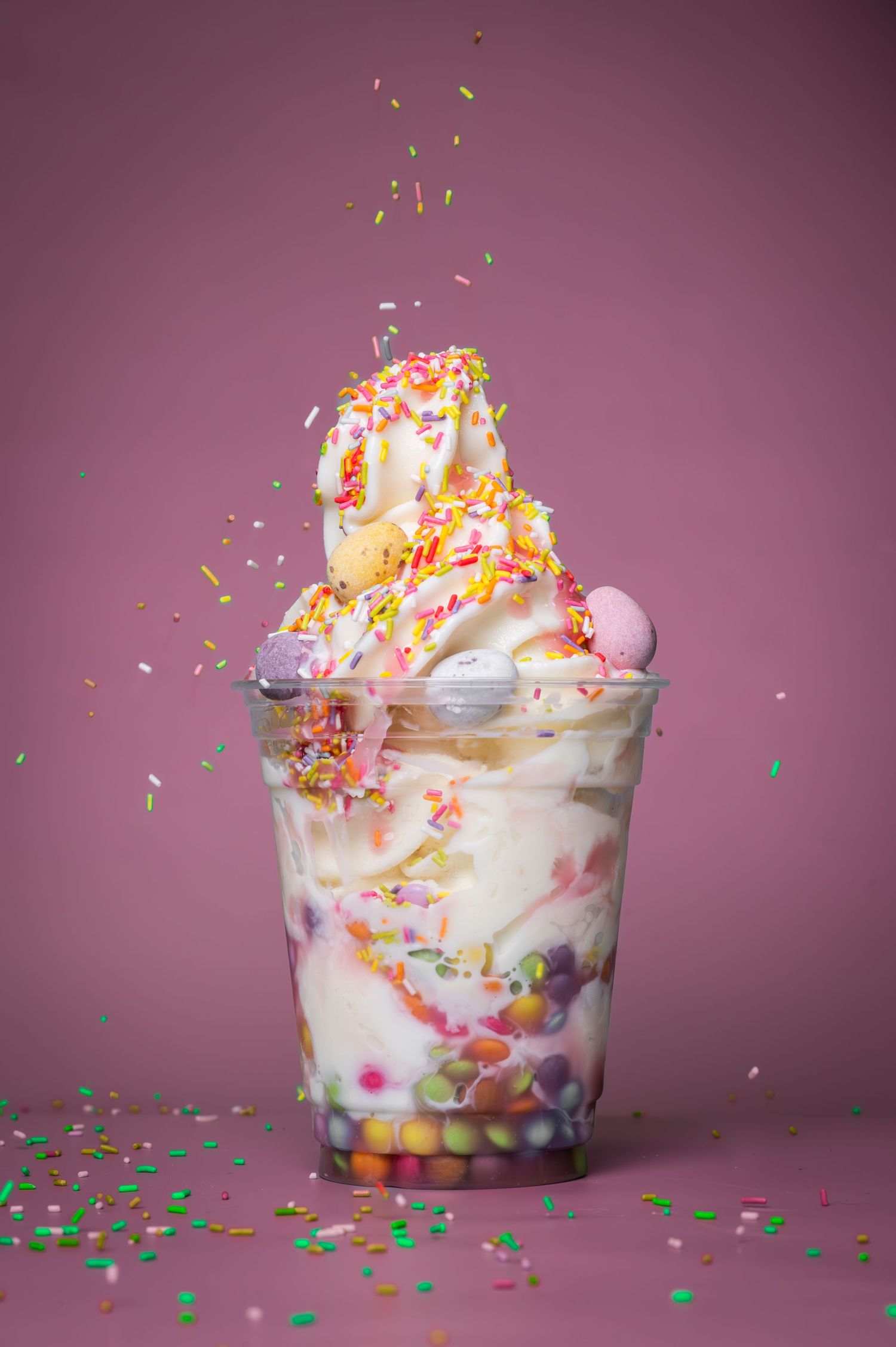 Colorful ice cream sundae in a plastic cup topped with sprinkles and candy on a pink background.