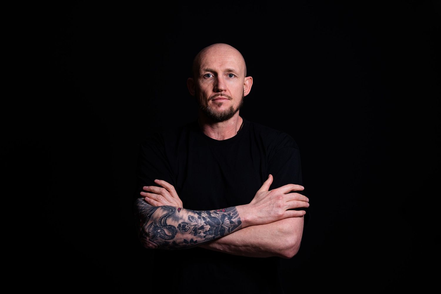 Tattooed man in black t-shirt standing with arms crossed against dark background.