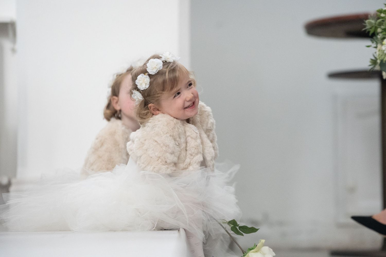 Two adorable flower girls in white tulle dresses with floral crowns sitting together at a wedding ceremony.