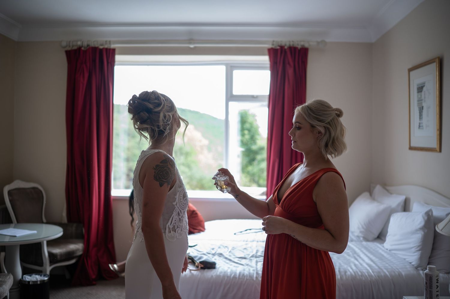 Bride in white wedding dress being helped by bridesmaid in red dress in a hotel room before the ceremony.