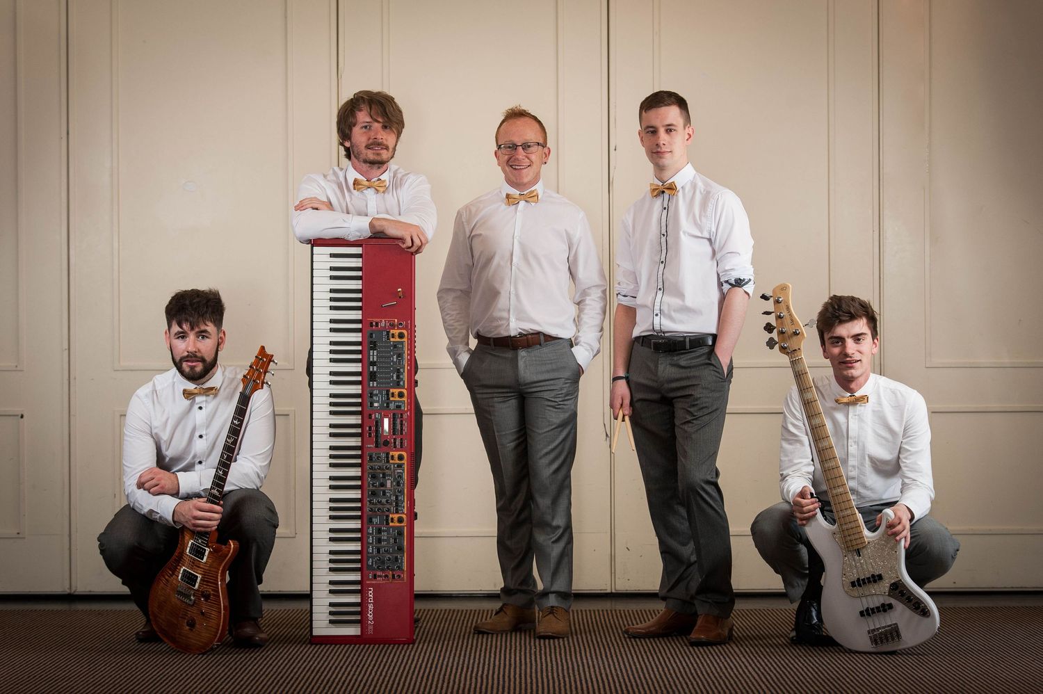 Five-piece band posing with instruments including keyboards and guitars in a bright room.