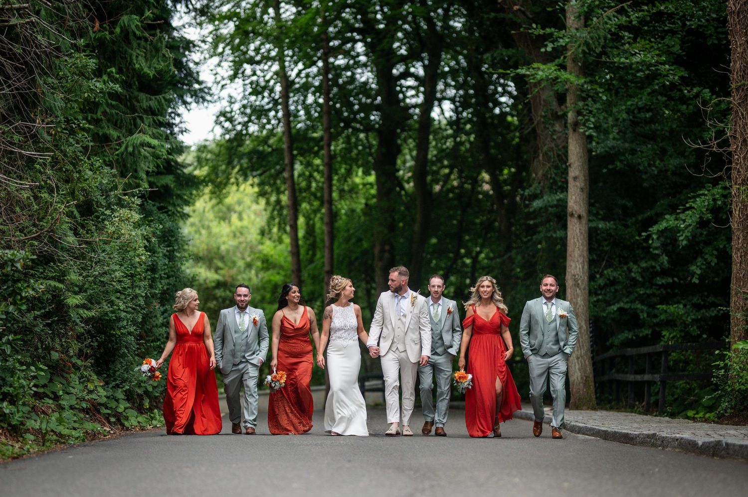 Wedding party walking down tree-lined road, bridesmaids in red dresses, groomsmen in grey suits.