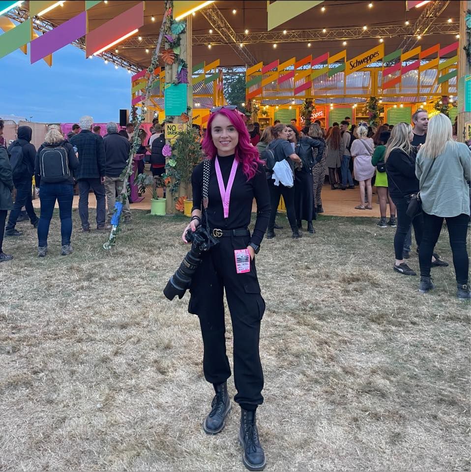Female photographer with pink hair holding camera at colorful outdoor festival event.