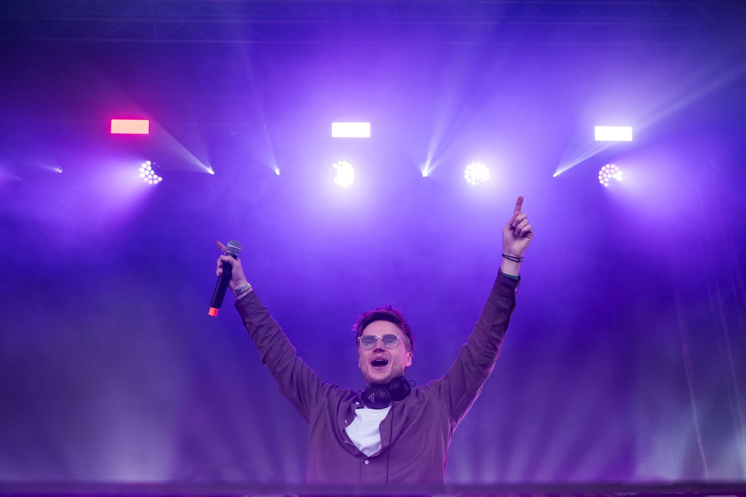 Male performer on stage with arms raised holding microphone under purple stage lighting.