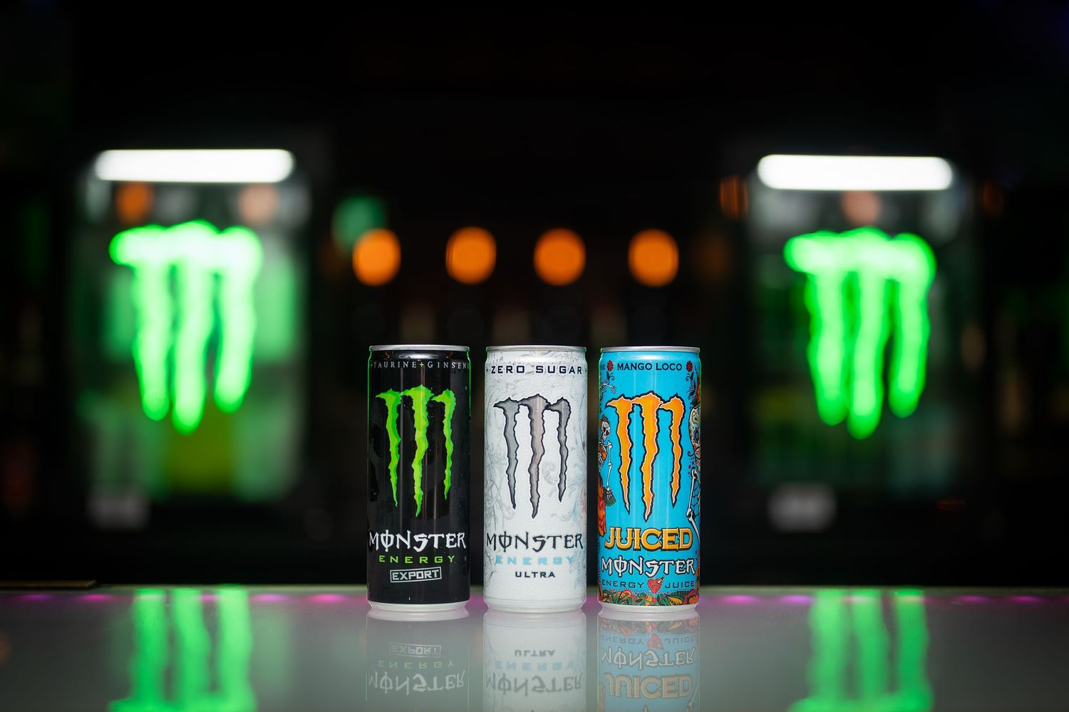 Three Monster Energy drink cans displayed on a reflective surface with green neon lights in background.