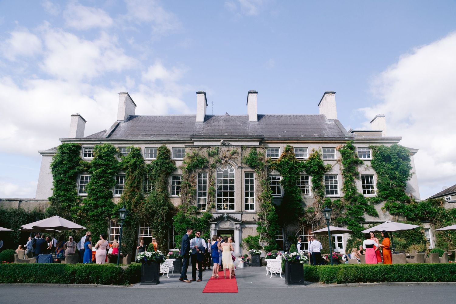 Elegant ivy-covered stone manor house wedding venue with red carpet entrance and guests gathered outside under blue skies.