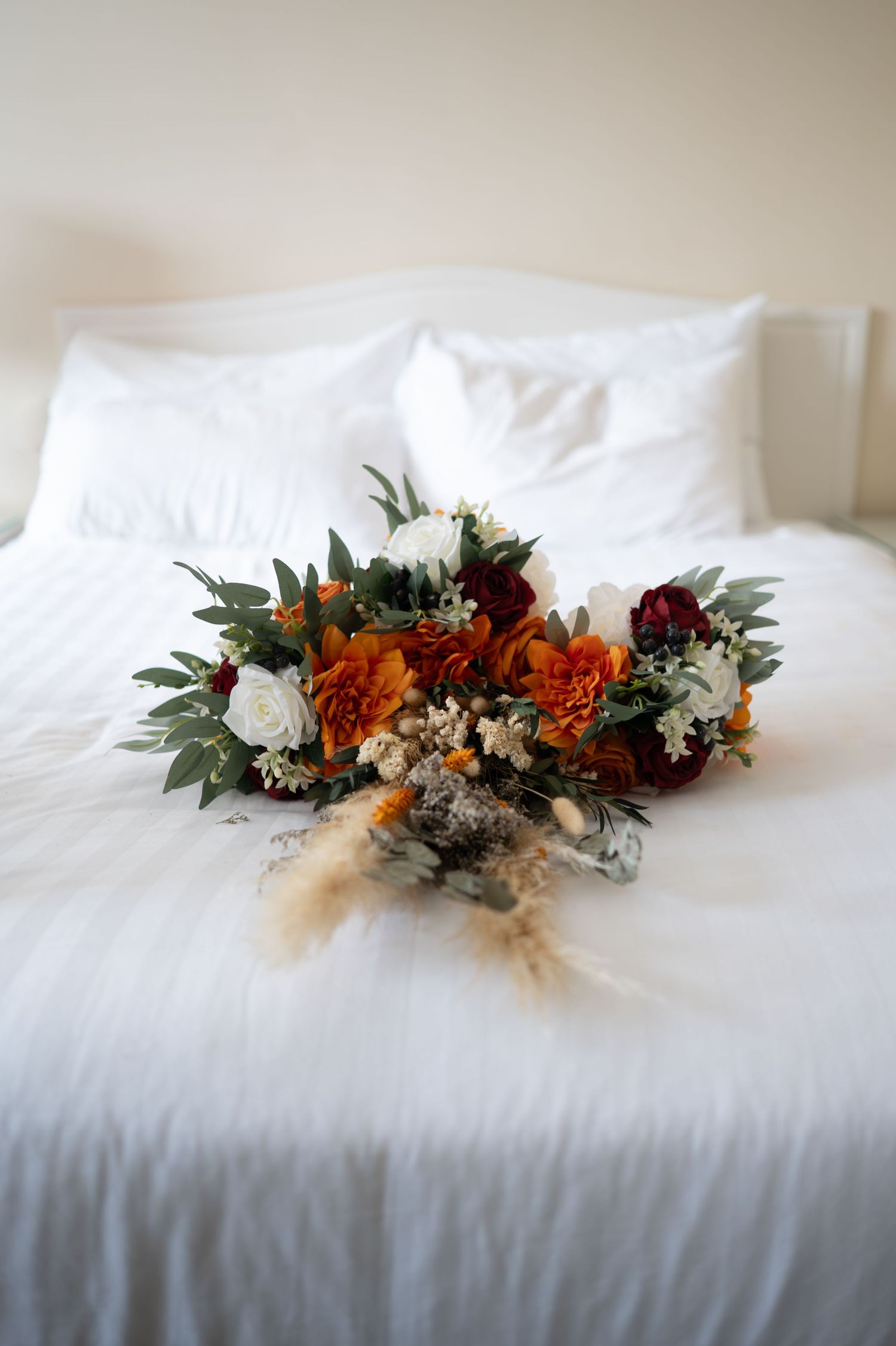 Rustic wedding bouquet with orange, burgundy, and white flowers on a white bed.