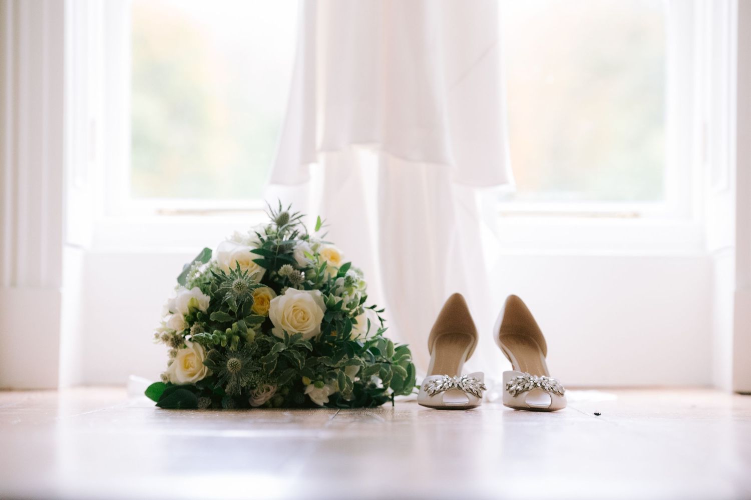 Bridal bouquet with white roses and greenery beside embellished heels on white floor near window.