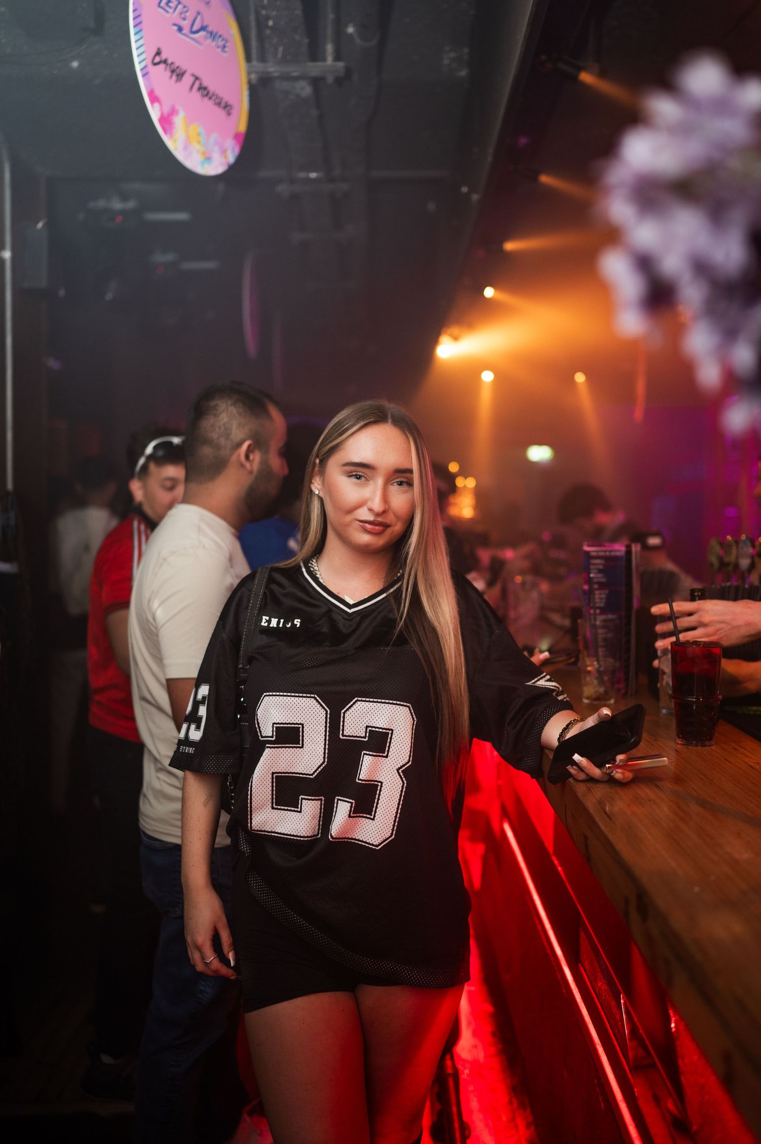 Young woman in a number 23 jersey posing at a nightclub bar with colorful lights.