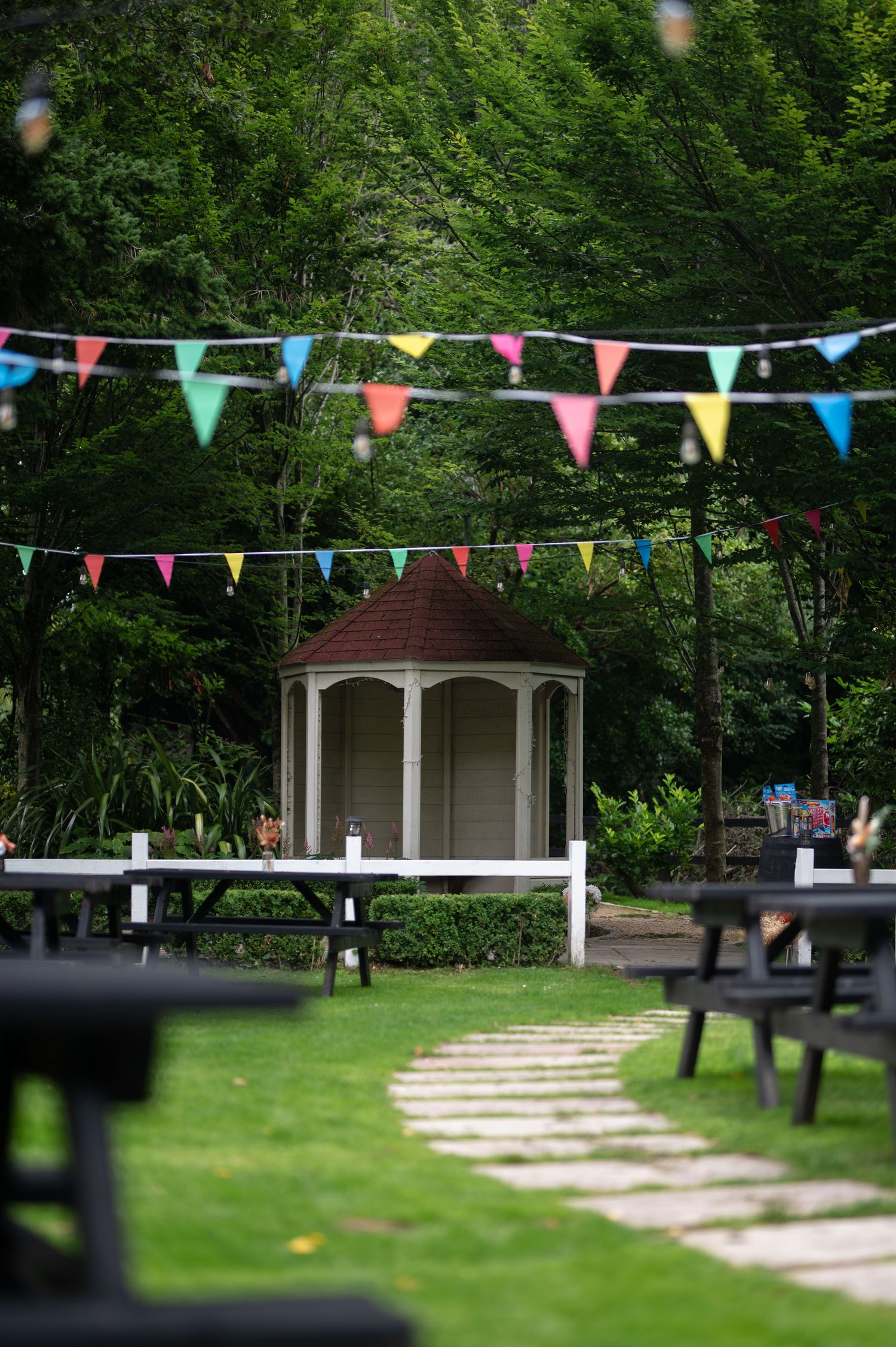 Colorful bunting flags hang over a lush garden with a white gazebo and stone pathway surrounded by picnic tables.