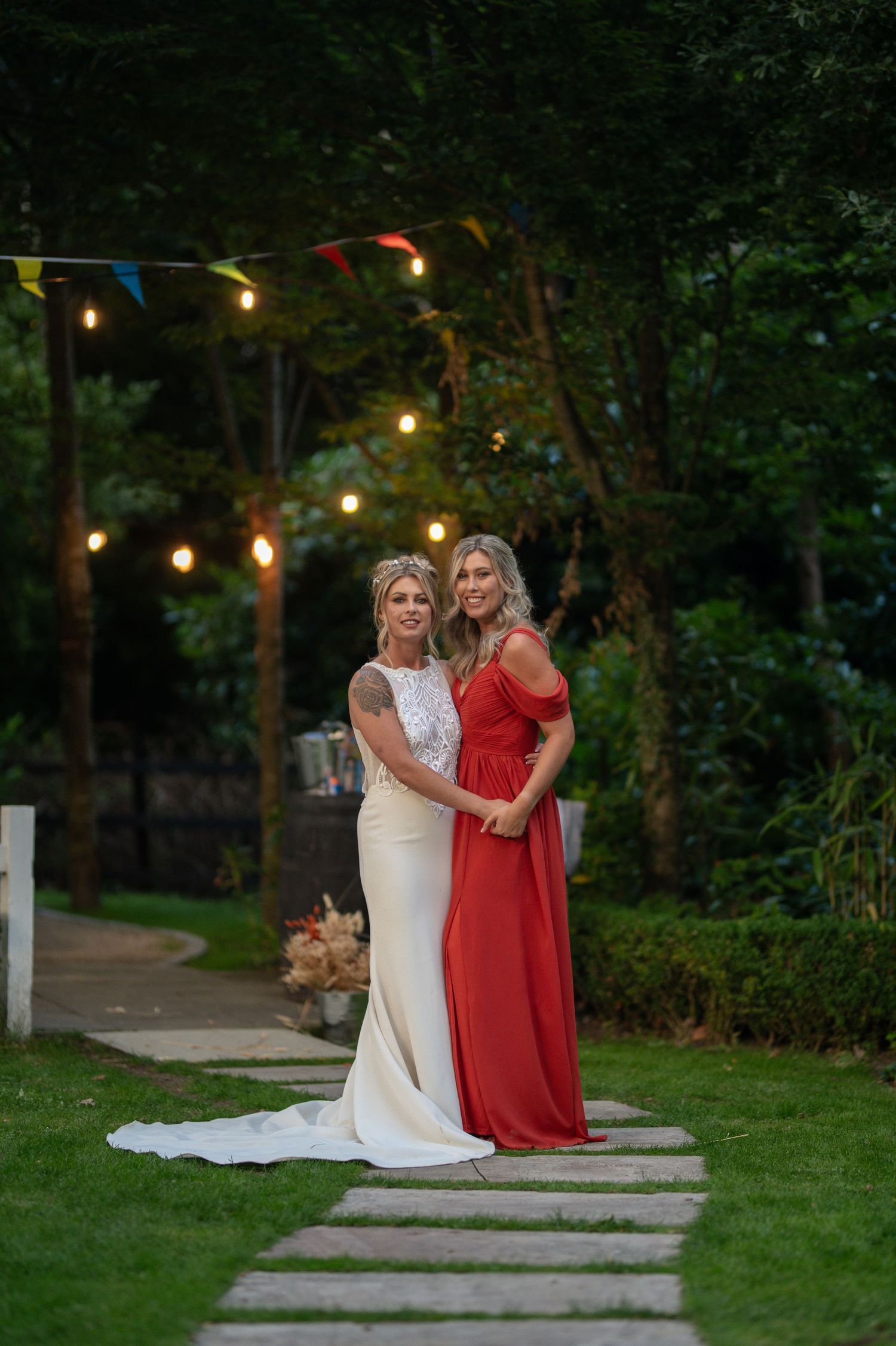 Bride in white gown and bridesmaid in red dress pose on garden path with string lights at dusk.