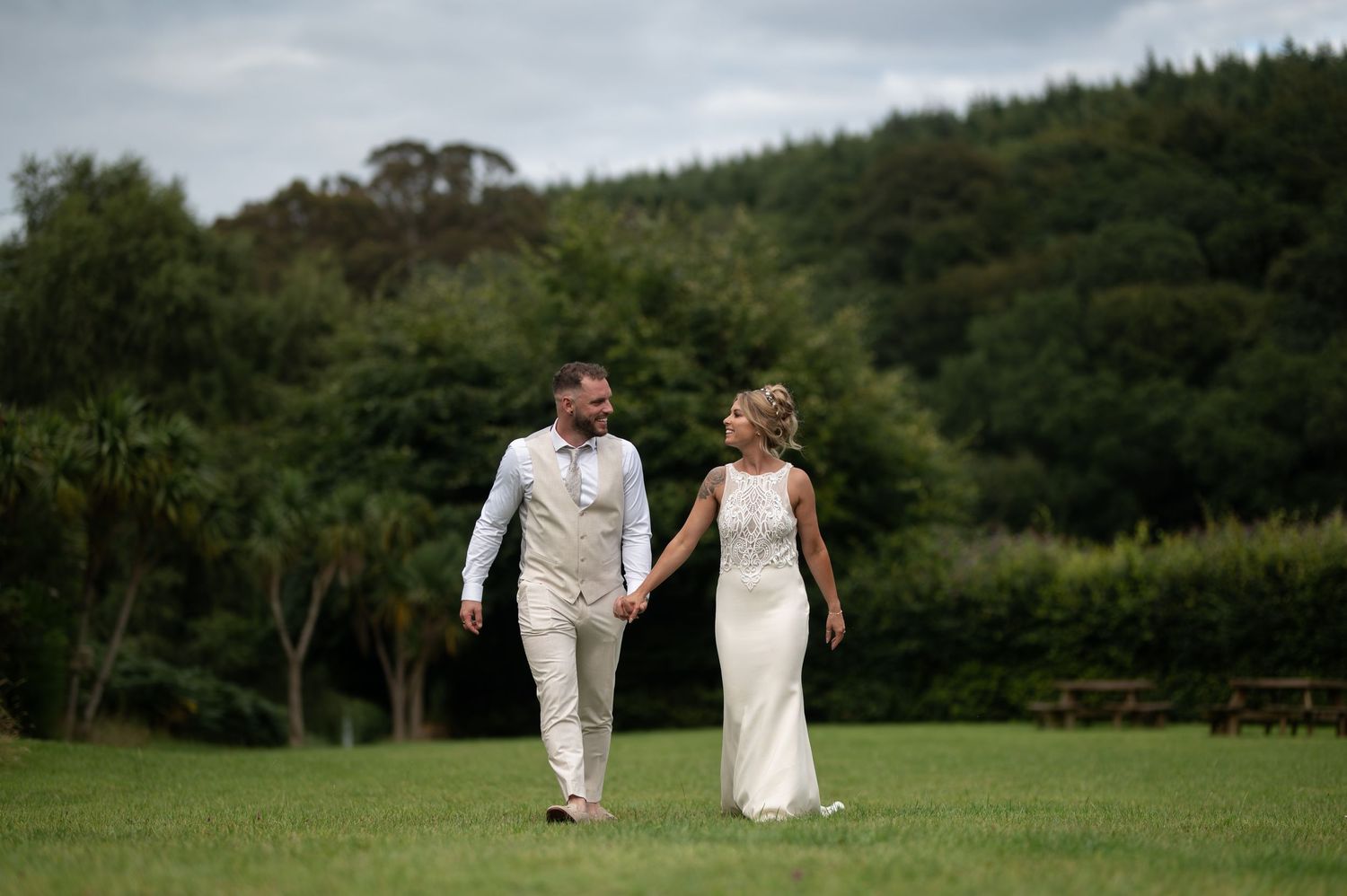 Bride and groom holding hands walking across green meadow with rolling hills in background.