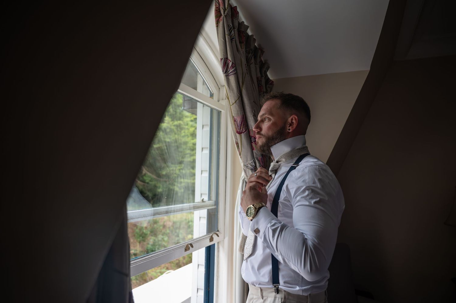 Groom adjusting his tie by a window in white shirt and suspenders, preparing for his wedding day.