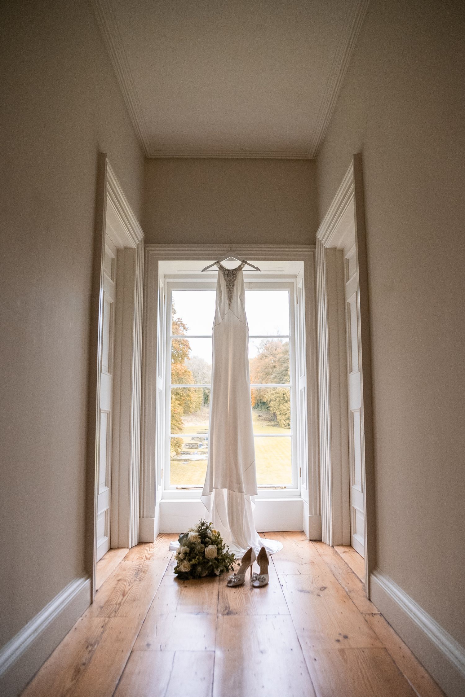 Wedding dress hanging in a sunlit hallway window with bridal bouquet and heels on hardwood floor.
