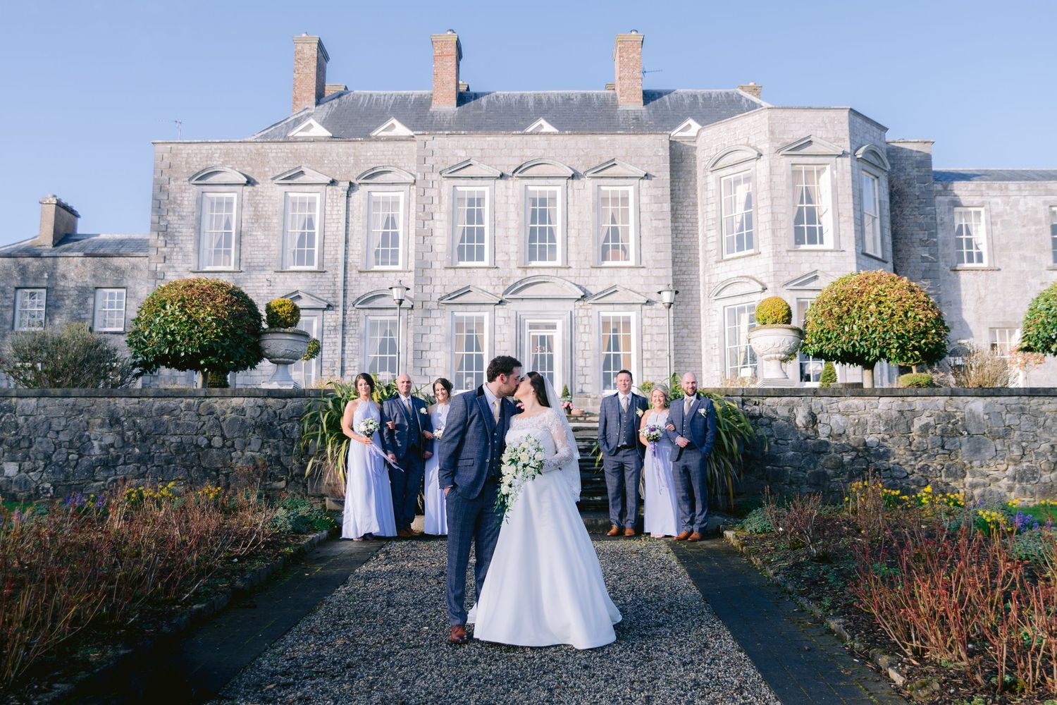 Bride and groom kissing in front of an elegant stone manor house with wedding party in lavender attire.