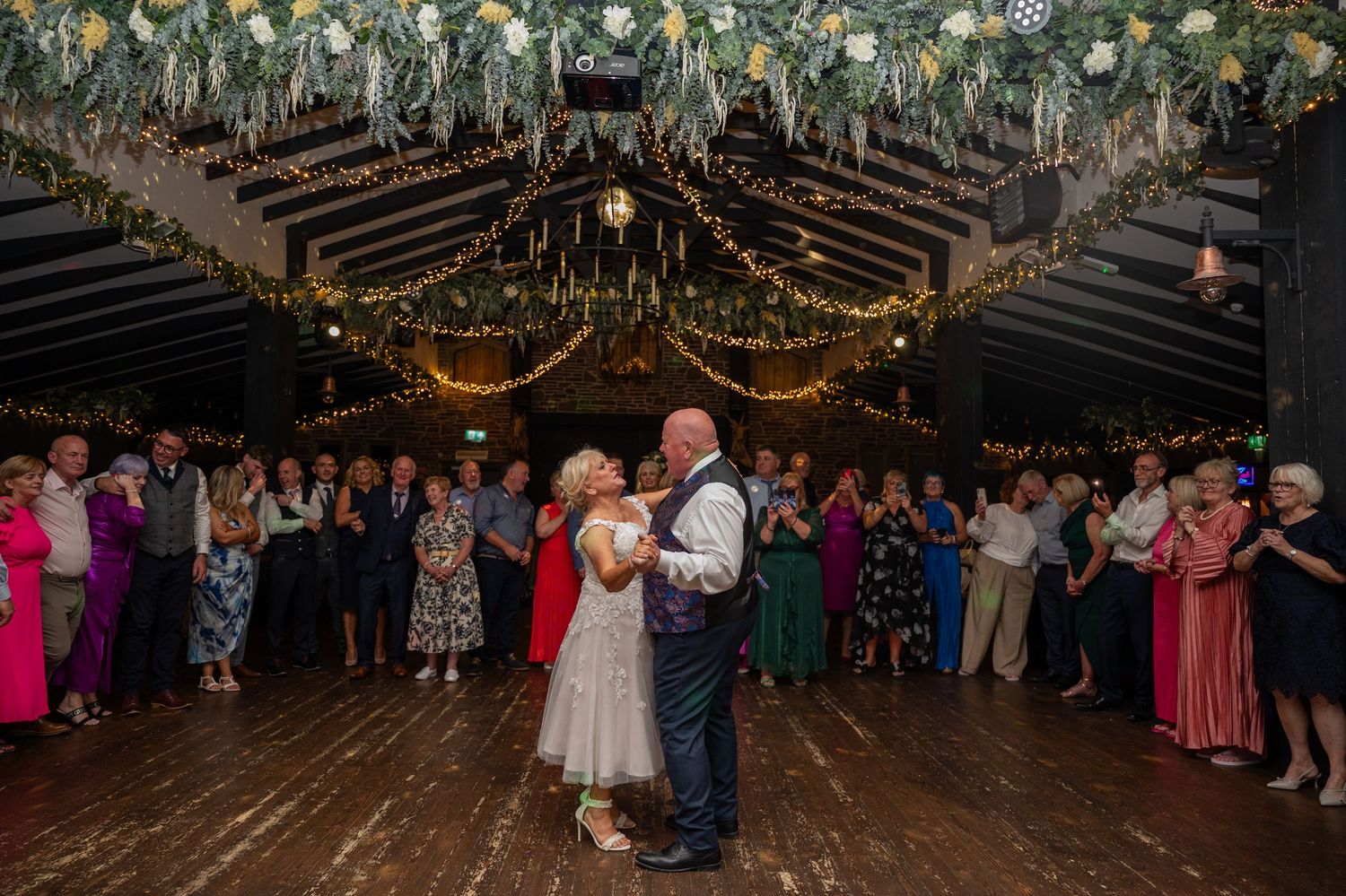 Couple sharing first dance in decorated barn venue with floral ceiling, fairy lights, and wedding guests watching.