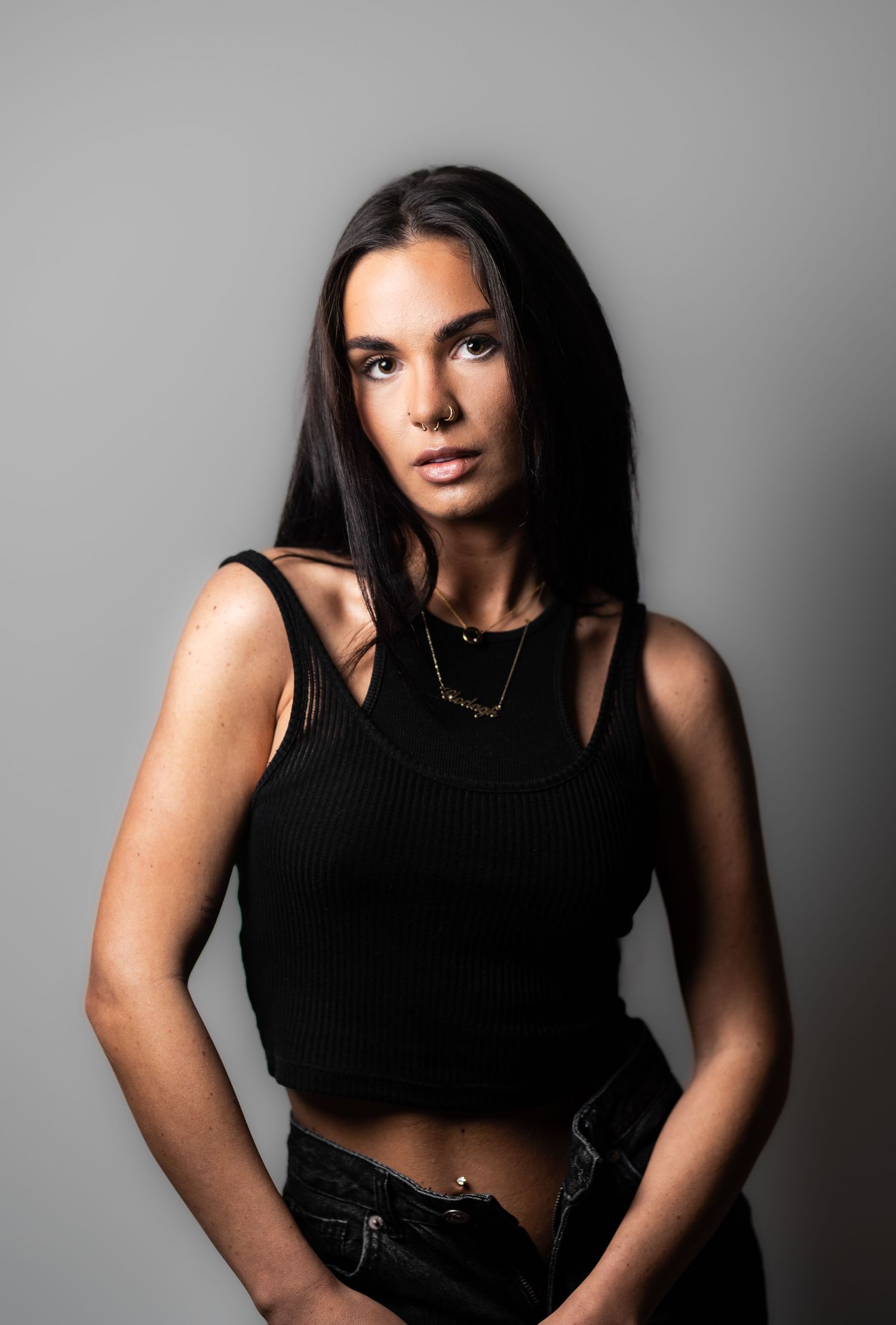 Young brunette woman in black crop top and jeans posing against grey studio background.