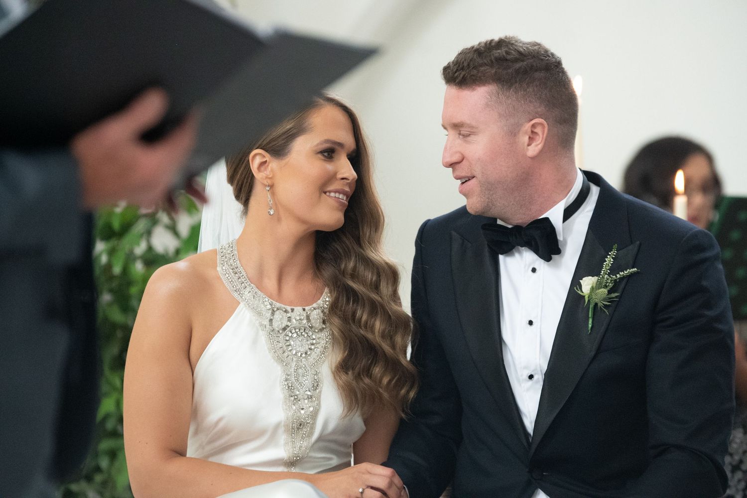 Bride and groom smiling at each other during wedding ceremony, bride in white gown, groom in black tuxedo.