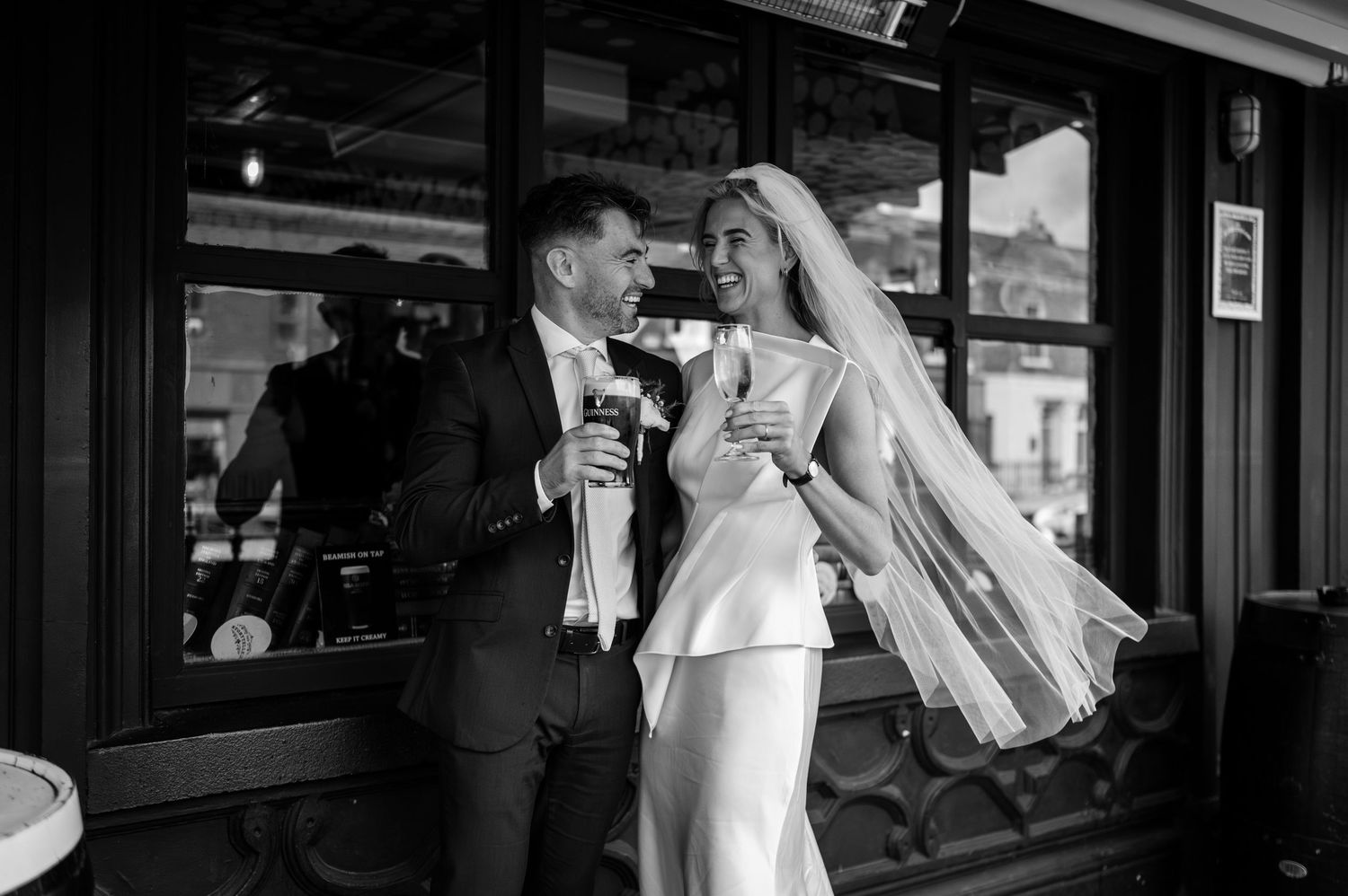 Black and white photo of a bride and groom laughing and holding drinks outside a venue on their wedding day.
