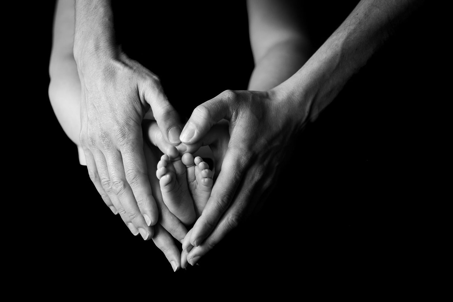 Two adult hands form a heart shape cradling tiny newborn baby feet in black and white.