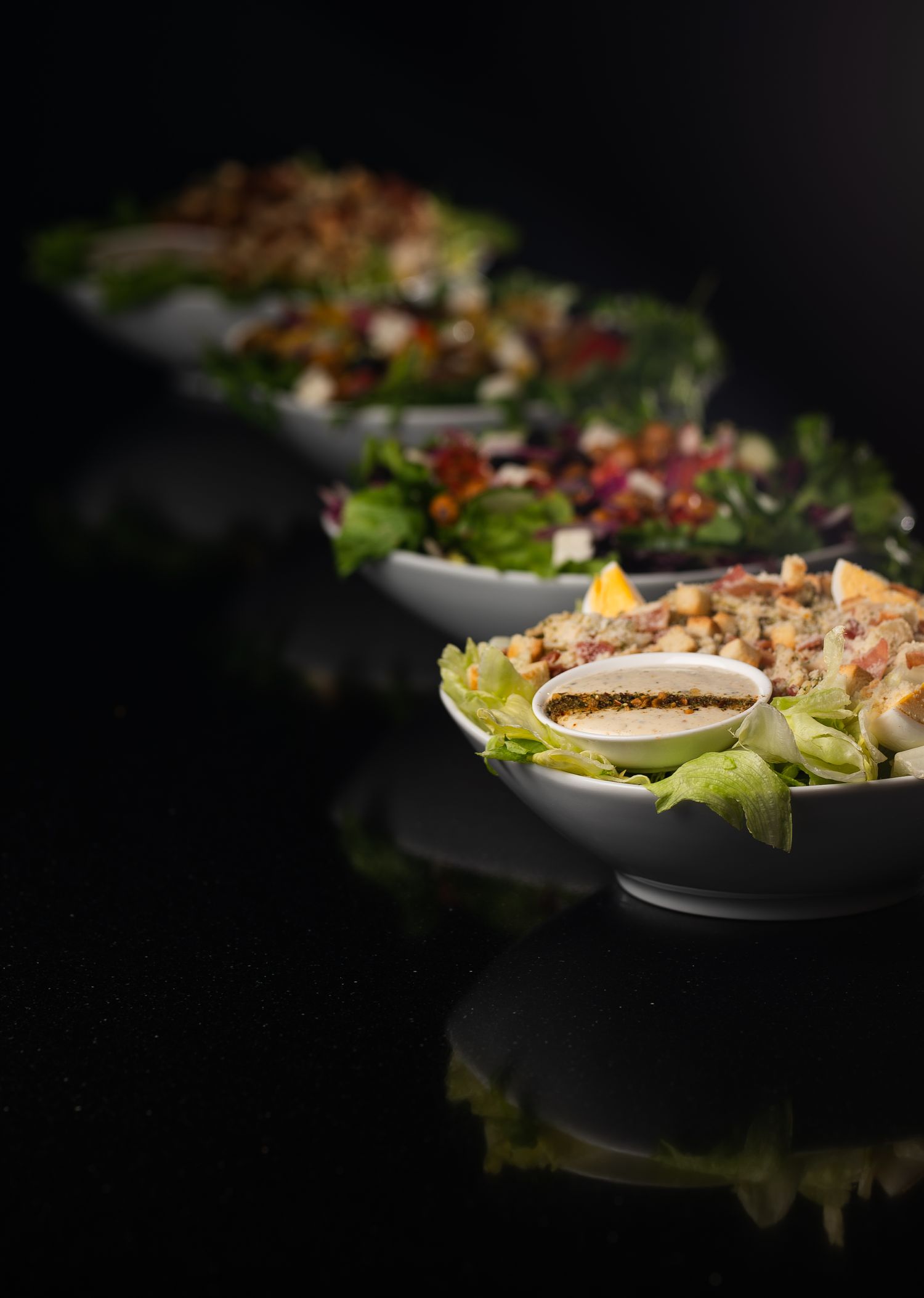 Three white bowls of fresh gourmet salads with dressing displayed on a dark background.