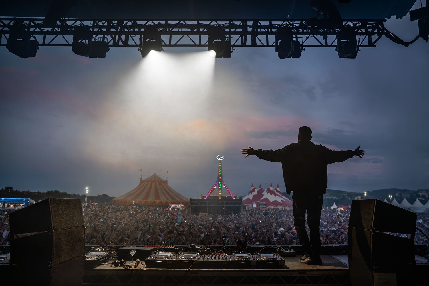 Silhouette of a DJ with arms outstretched facing a massive festival crowd at dusk.
