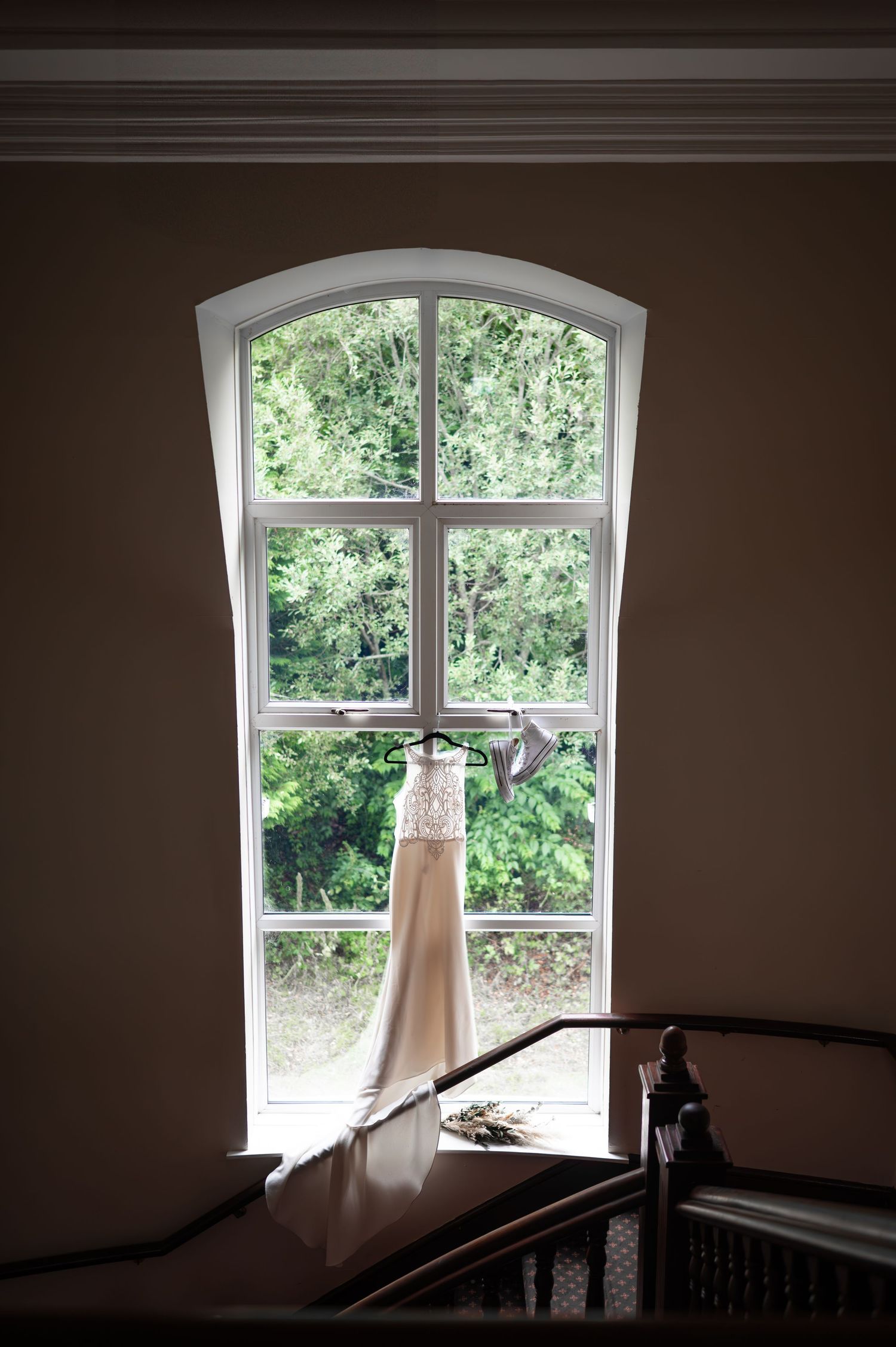 Wedding dress hanging in an arched window with lush green trees visible outside in natural light.