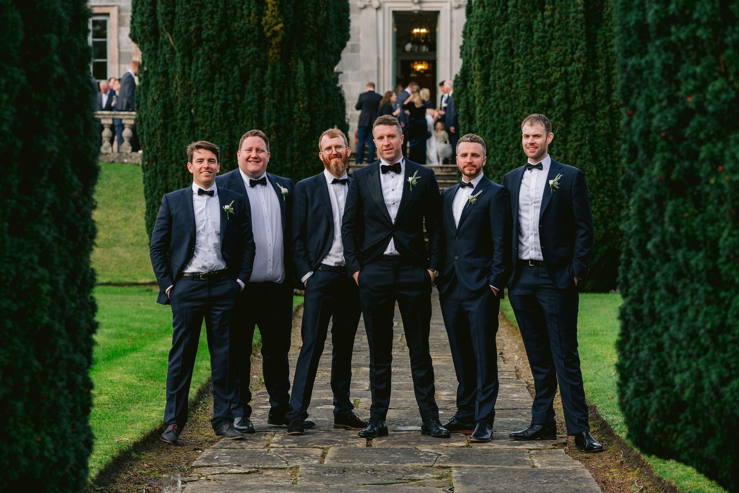 Six groomsmen in black tuxedos with bow ties posing outdoors between tall hedges at a wedding venue.