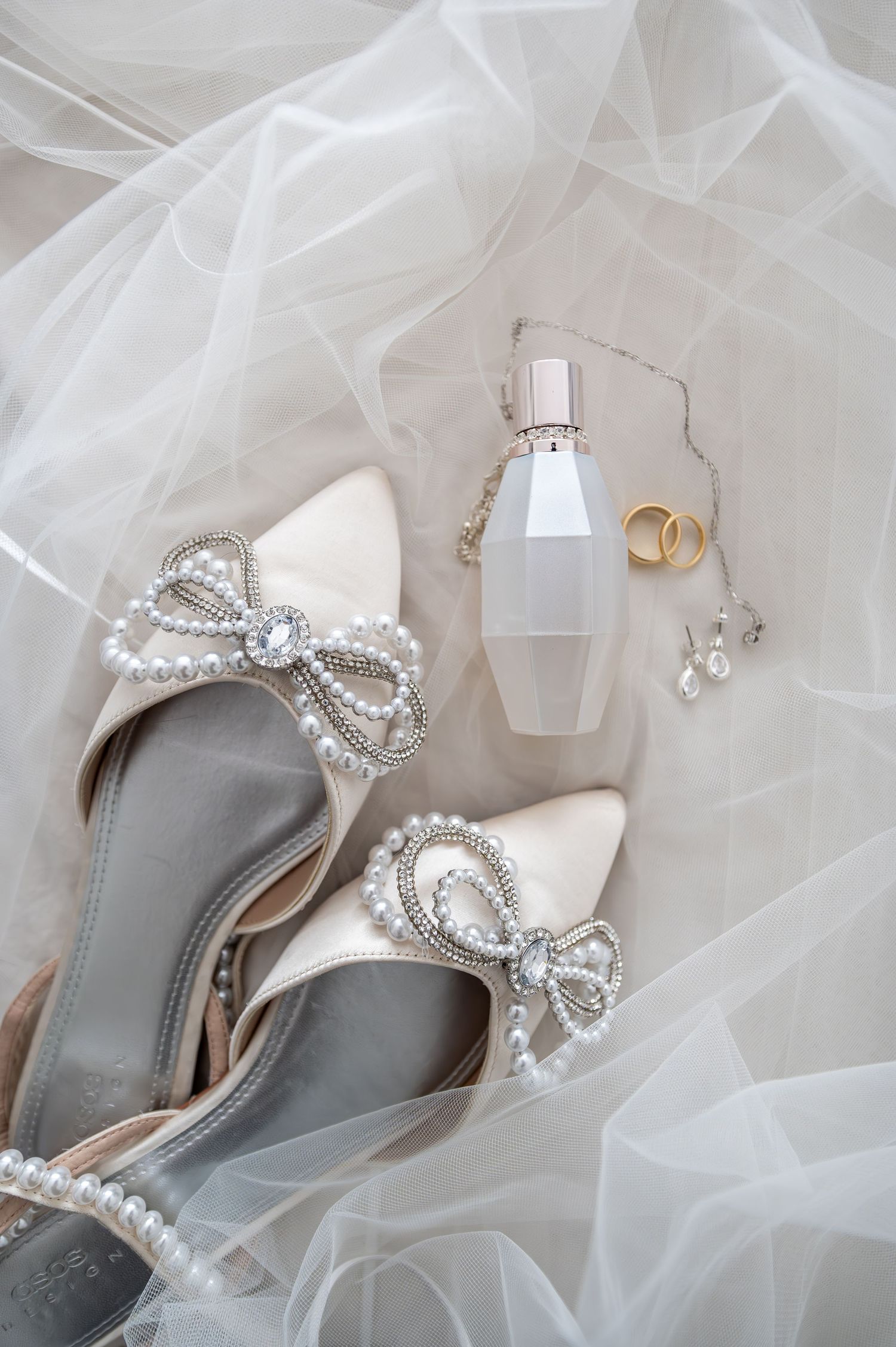 Elegant bridal flat lay with pearl bow heels, perfume bottle, gold rings, and delicate earrings on white tulle veil.