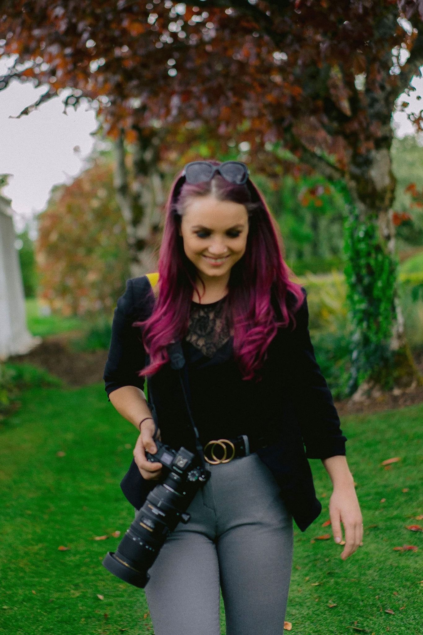 Female photographer with purple hair holding DSLR camera in autumn garden setting.