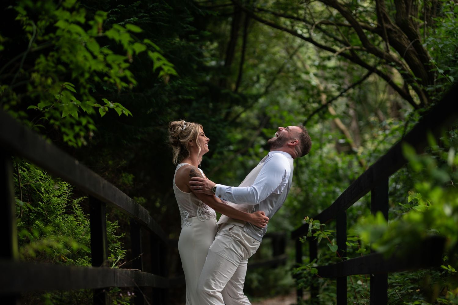 Bride and groom sharing joyful embrace on wooden bridge surrounded by lush green forest.