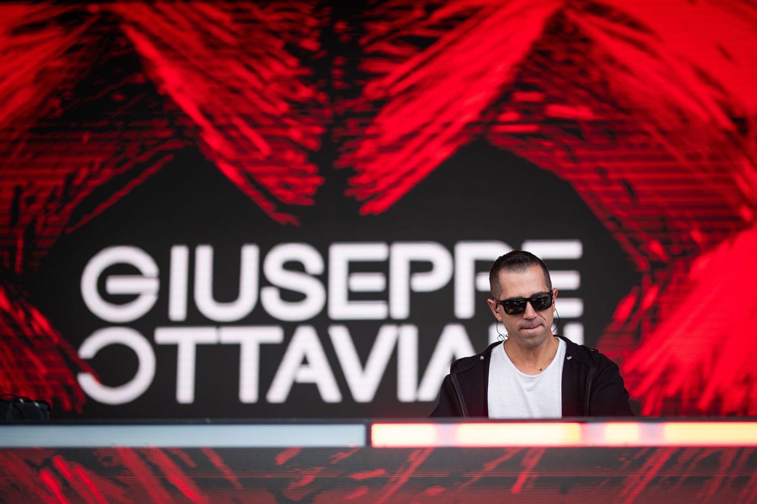 DJ Giuseppe Ottaviani performing behind decks with red backdrop signage at a music festival.