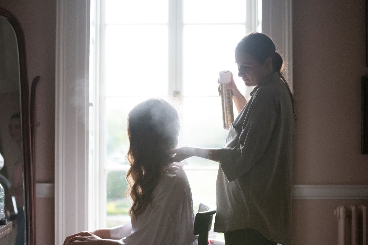 Woman styling another woman's hair by a bright window while holding a cup, in a softly lit elegant room.