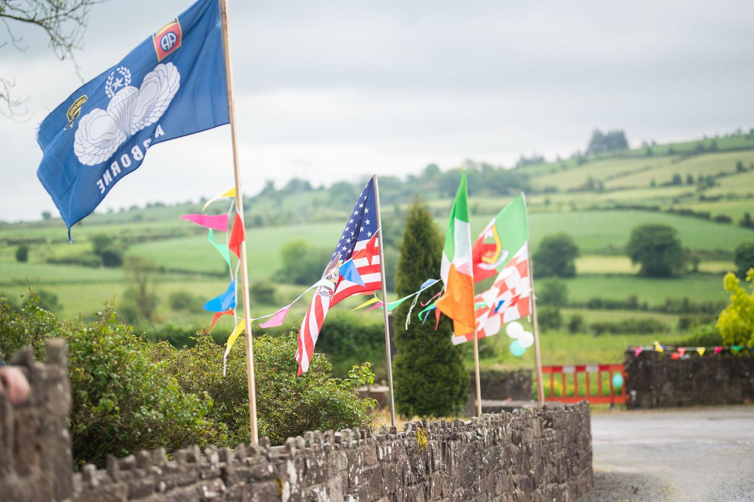 Colorful international flags including USA, Wales, and Ireland flying on poles beside a stone wall with green hills.