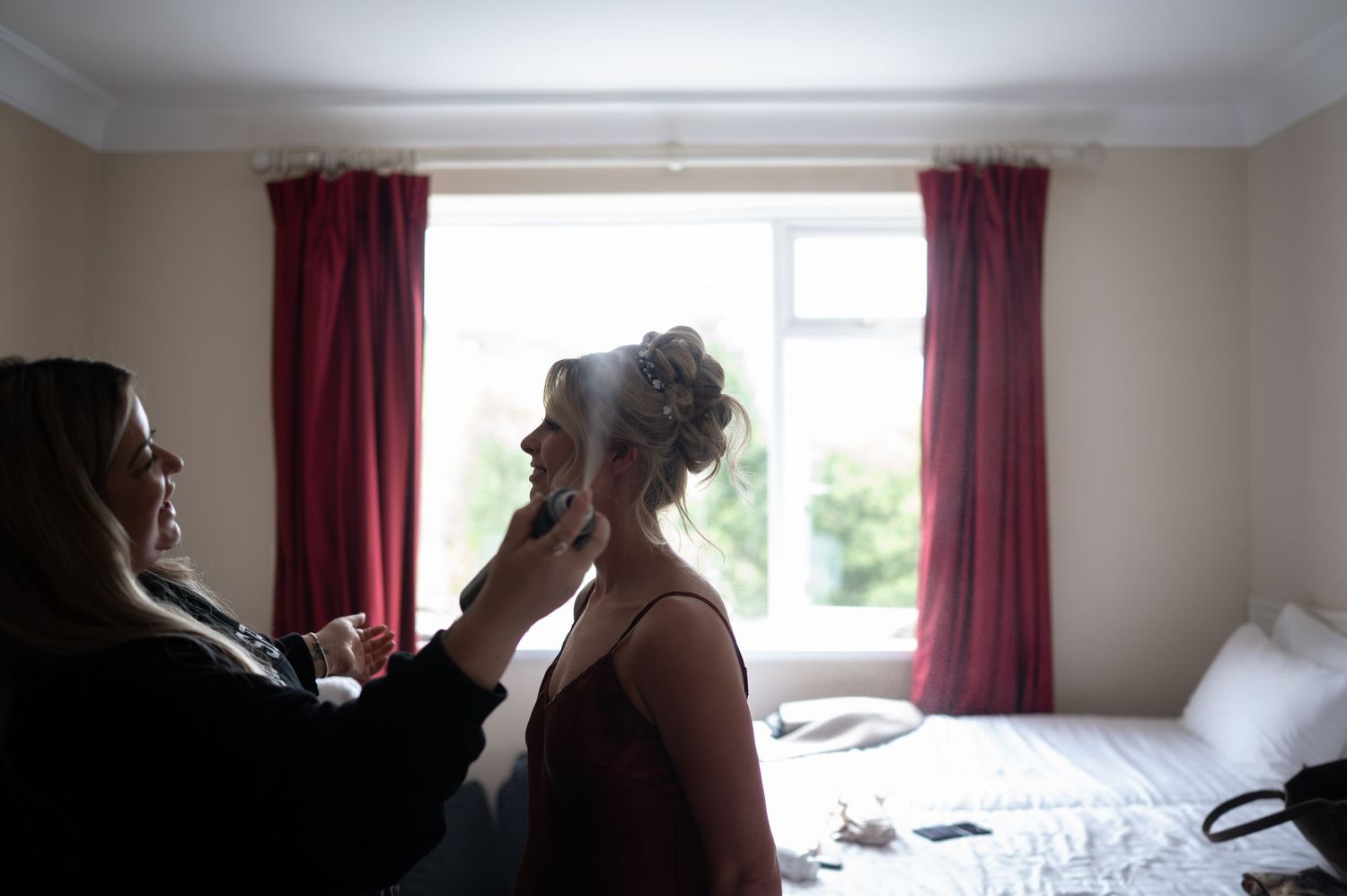 Bridesmaid adjusting bride's veil in a softly lit hotel room with red curtains before the wedding ceremony.